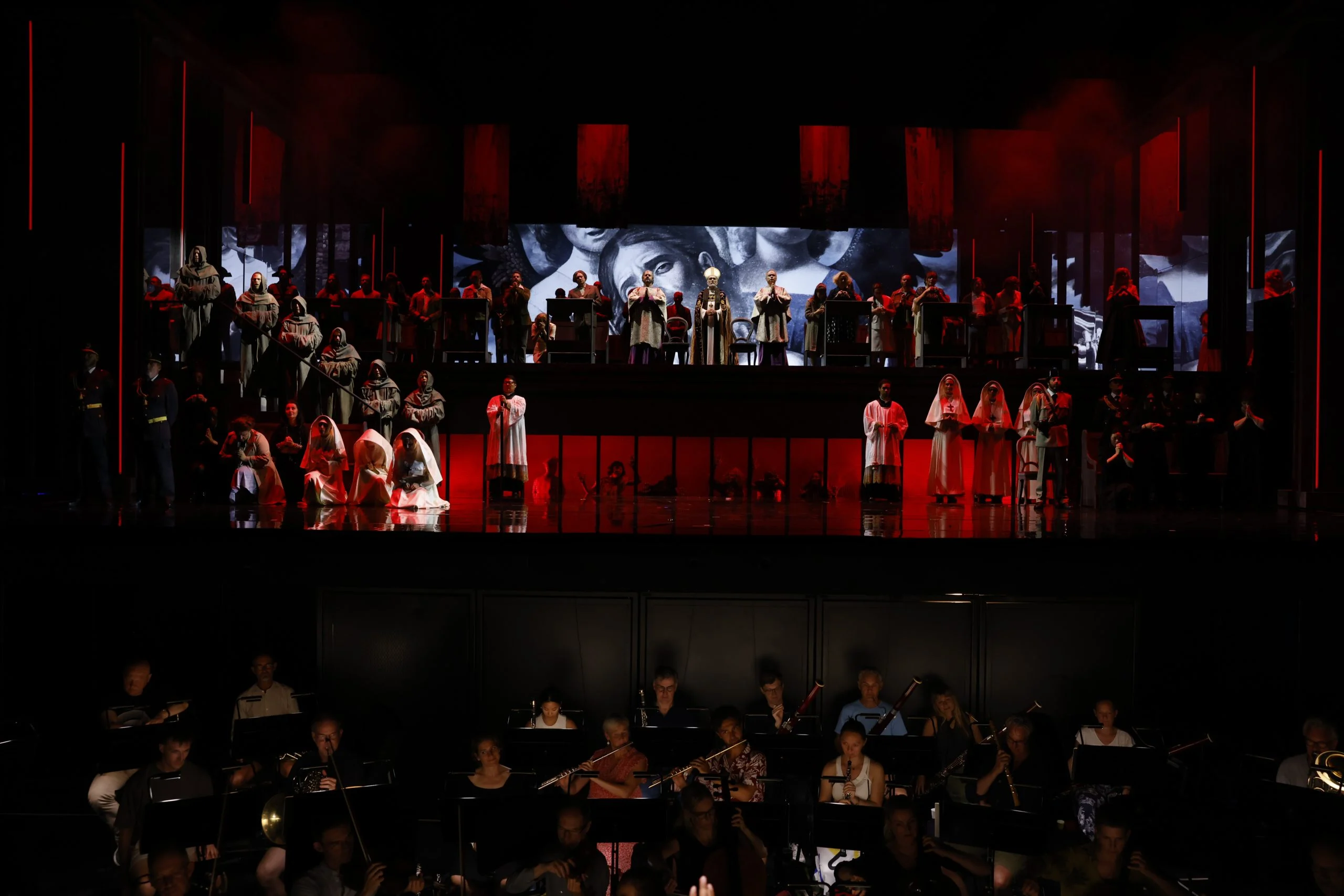 Hooded penitents and chorus on raised red-lit tiers above the orchestra pit, projected faces behind – Don Carlo at the Royal Danish Theatre, Copenhagen, Denmark, 2024 – opera set design