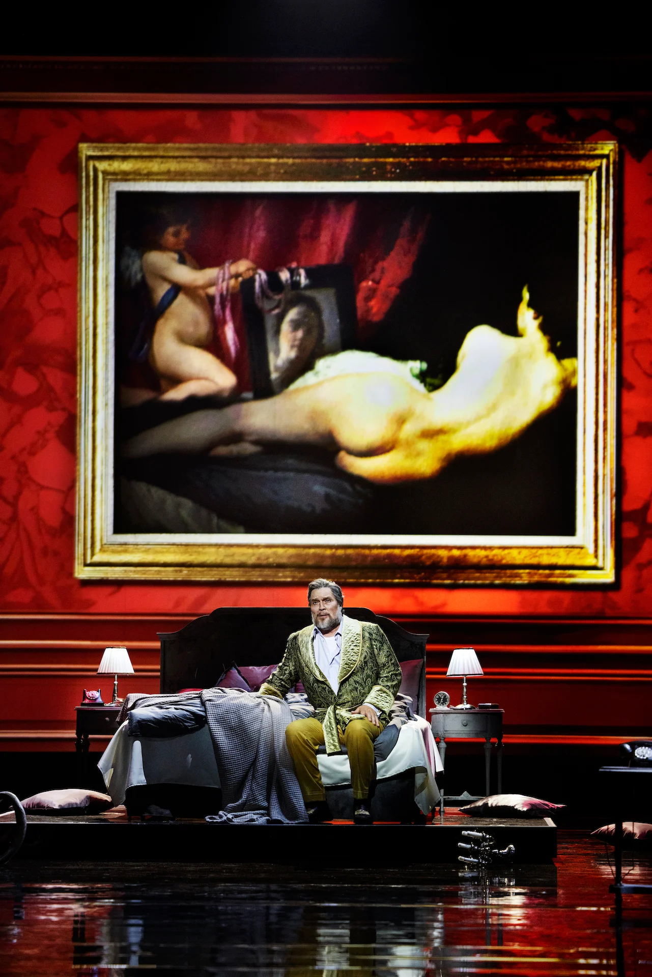 Singer in a green brocade robe seated on a bed beneath a giant framed painting of a reclining nude – Don Carlo at the Royal Danish Theatre, Copenhagen, Denmark, 2024 – opera set design