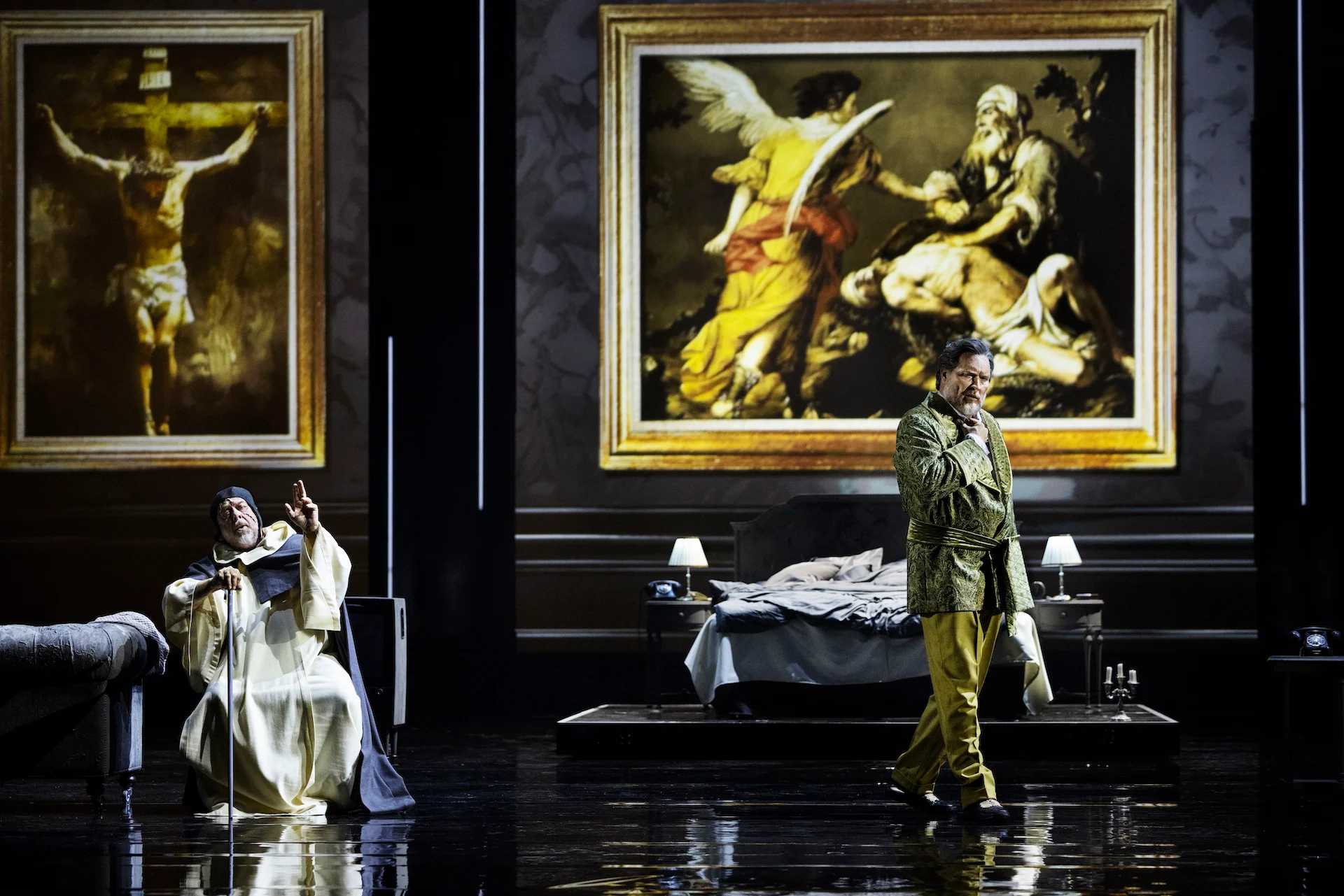 Bedroom scene with a cleric in white and a singer in a green robe, framed religious paintings behind – Don Carlo at the Royal Danish Theatre, Copenhagen, Denmark, 2024 – opera set design