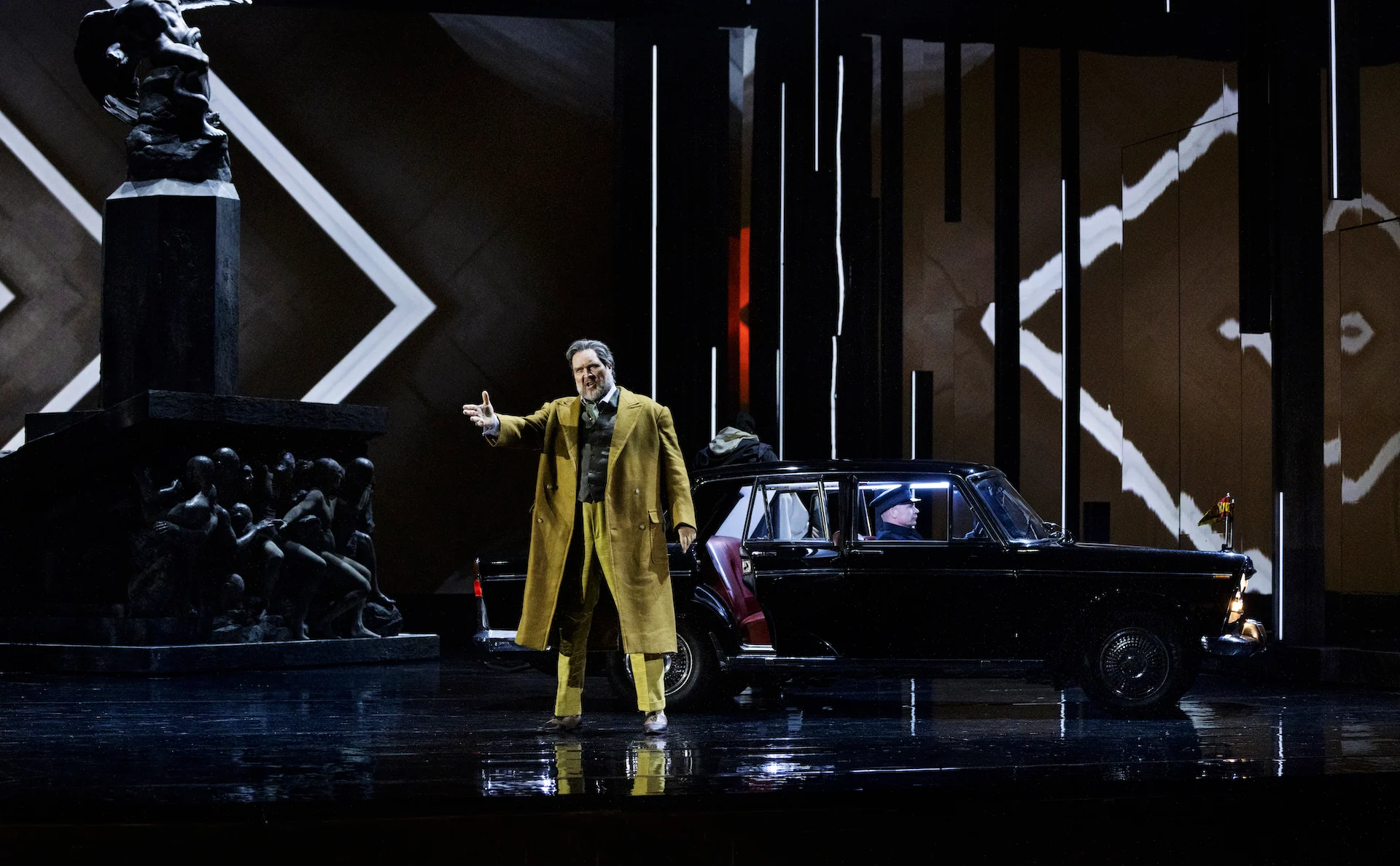 Singer in a yellow overcoat gesturing beside a black vintage car, the monument and white zigzag lines behind – Don Carlo at the Royal Danish Theatre, Copenhagen, Denmark, 2024 – opera set design