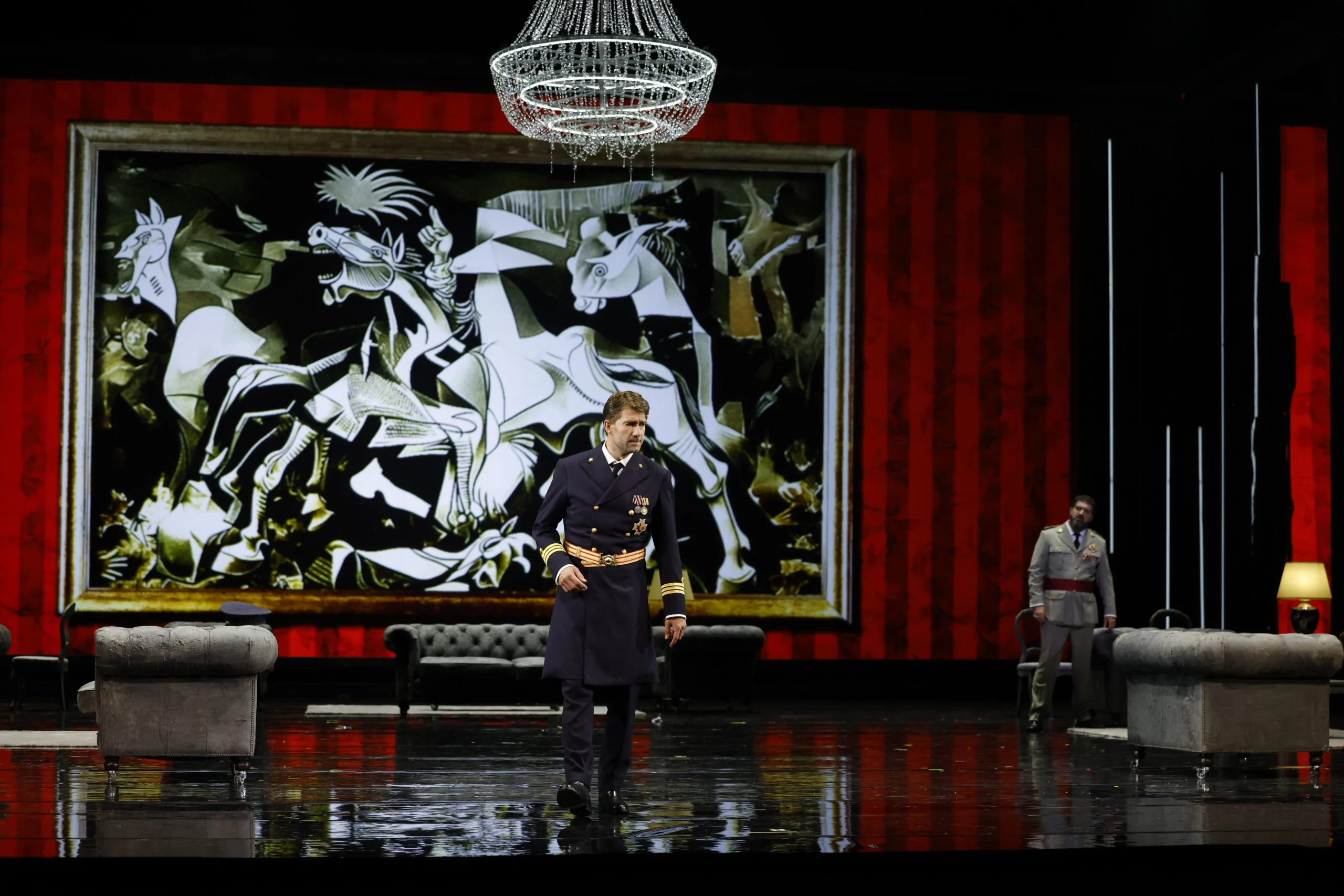 Officer in naval uniform before a huge reproduction of Picasso's Guernica, a chandelier and red curtains – Don Carlo at the Royal Danish Theatre, Copenhagen, Denmark, 2024 – opera set design