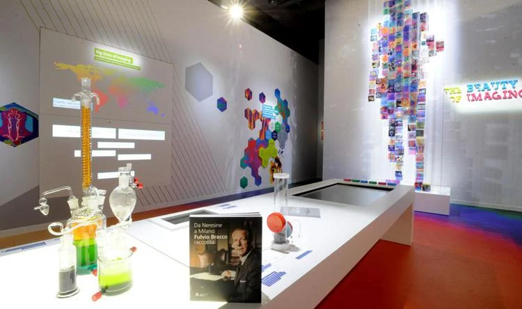 Exhibition room with lab glassware and a book on a white table, a figure of coloured images behind – Bracco The Beauty of Imaging, Milan, Italy, 2017 – exhibition design