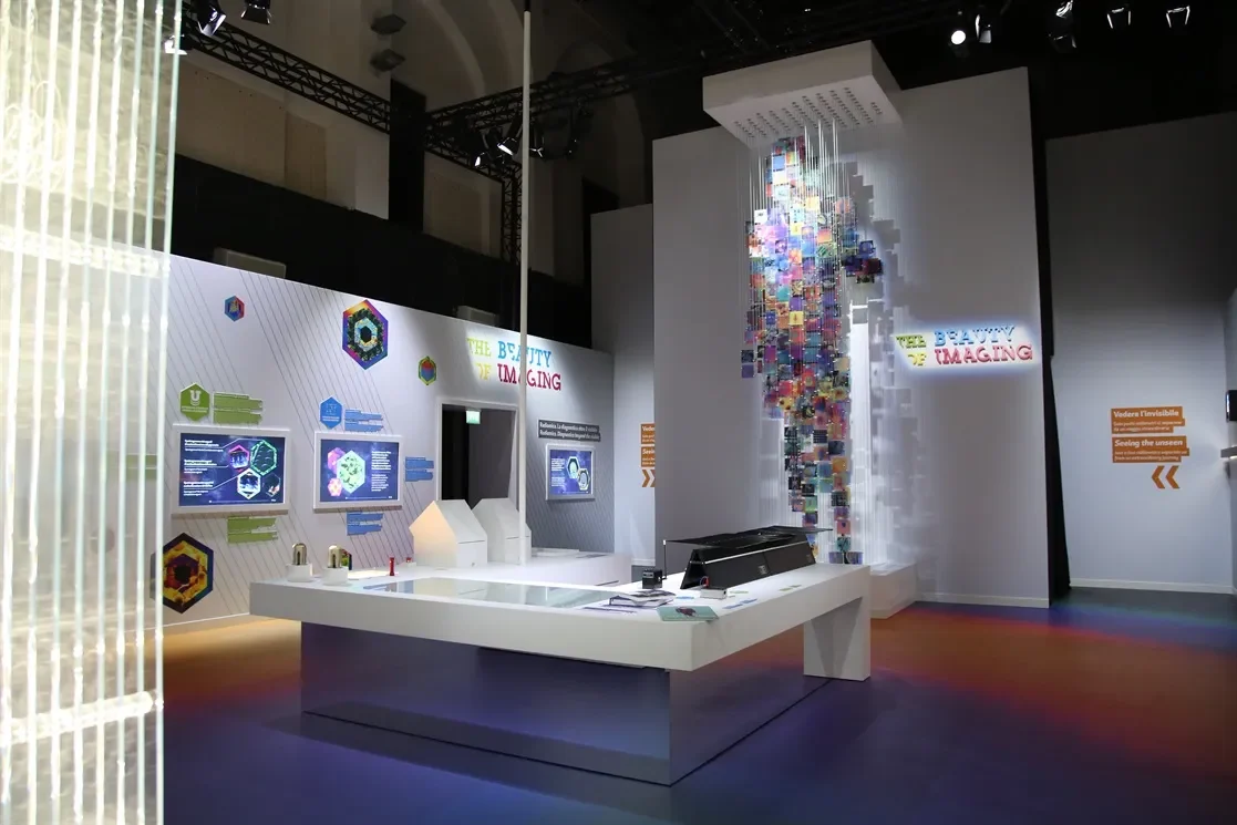 Wide view of the exhibition hall with a white central table, wall graphics and the hanging image figure – Bracco The Beauty of Imaging, Milan, Italy, 2017 – exhibition design