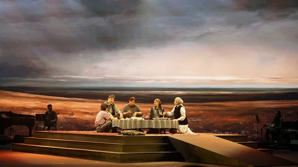 Opera scene: family seated at a table on a stepped platform before a vast steppe landscape projection – Waltz Karoly at Turkistan Theatre, Kazakhstan, 2024 – set design