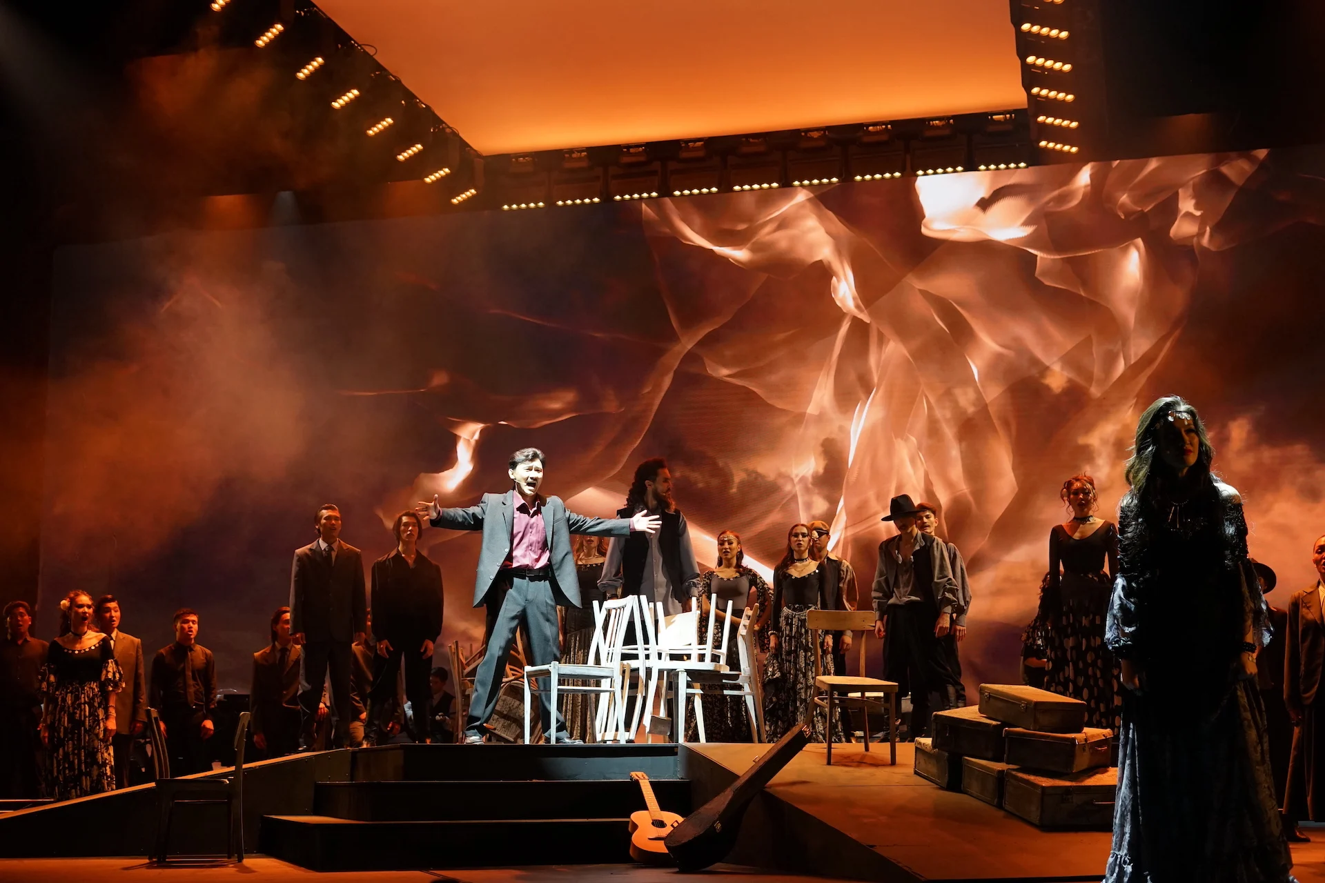 Ensemble scene with white chairs and stacked crates, a singer with open arms before swirling orange projections – Waltz Karoly at Turkistan Theatre, Kazakhstan, 2024 – set design