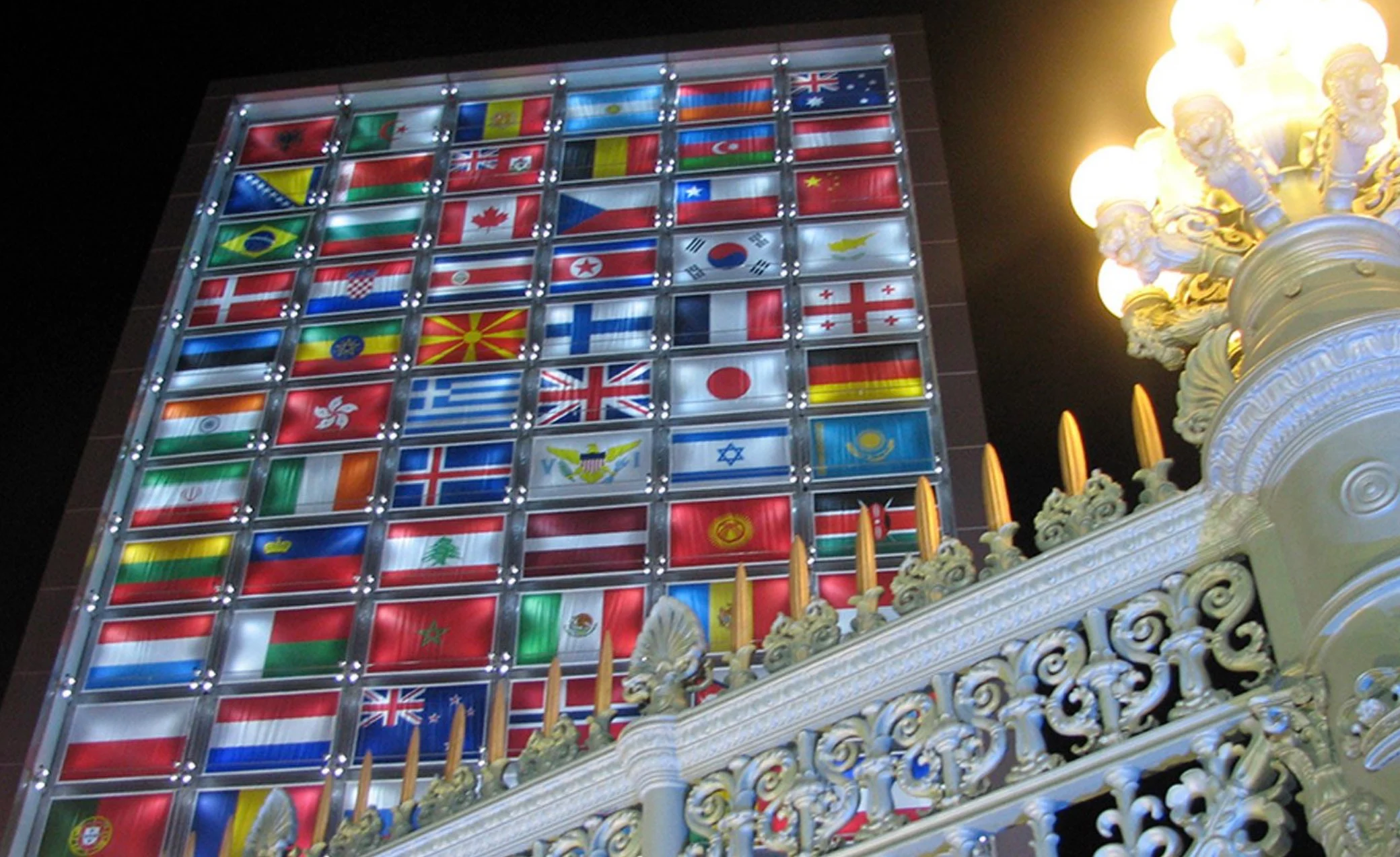 Illuminated wall of national flags at night, seen behind an ornate white balustrade and street lamp – Medals Plaza, Turin 2006 Winter Olympics, Italy – event design