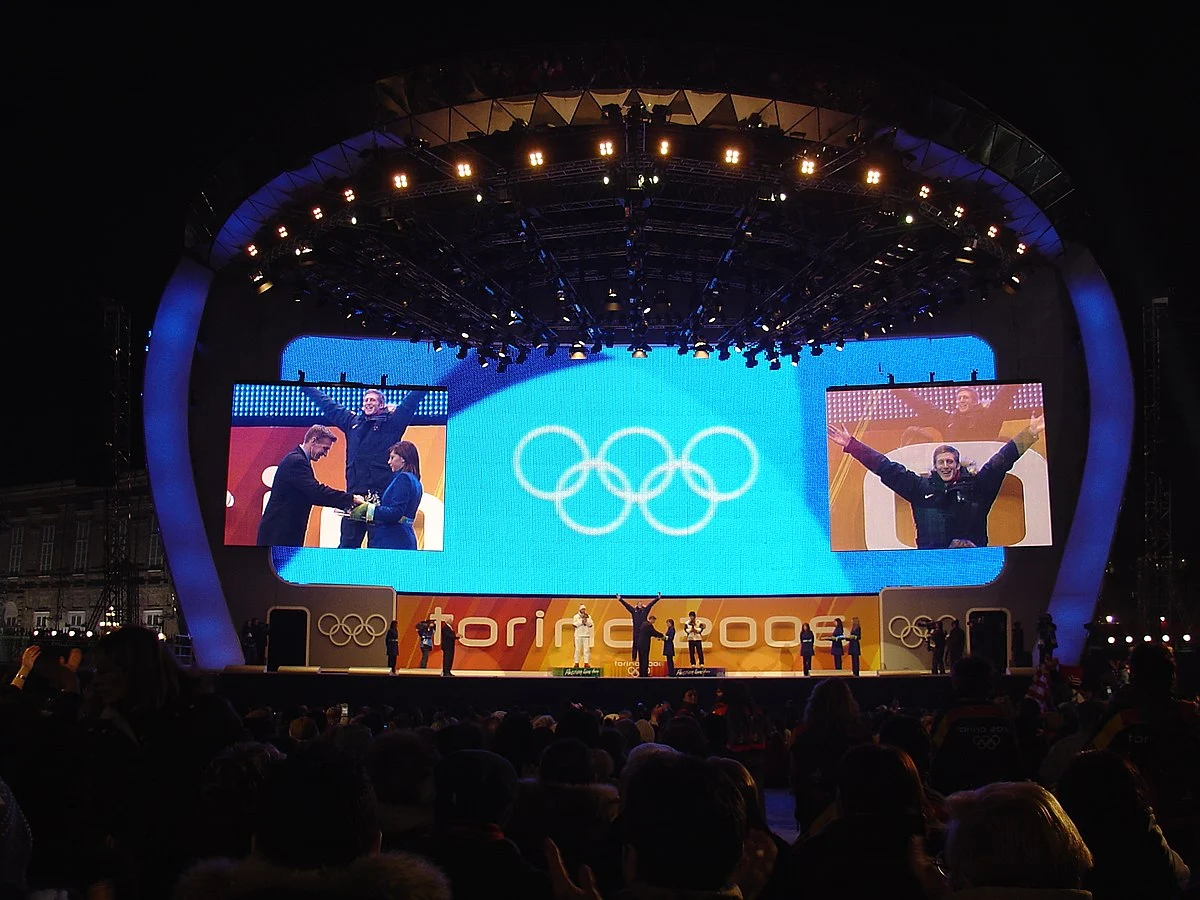 Medal ceremony on the curved stage, Olympic rings on the central screen and Torino 2006 along the front – Medals Plaza, Turin 2006 Winter Olympics, Italy – event design