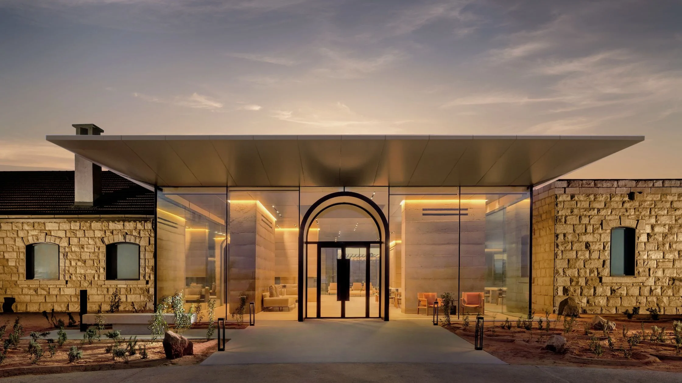 Hotel entrance at dusk: a glass pavilion under a thin flat roof between two old stone buildings – The Chedi Hegra, AlUla, Saudi Arabia, 2024 – hotel architecture