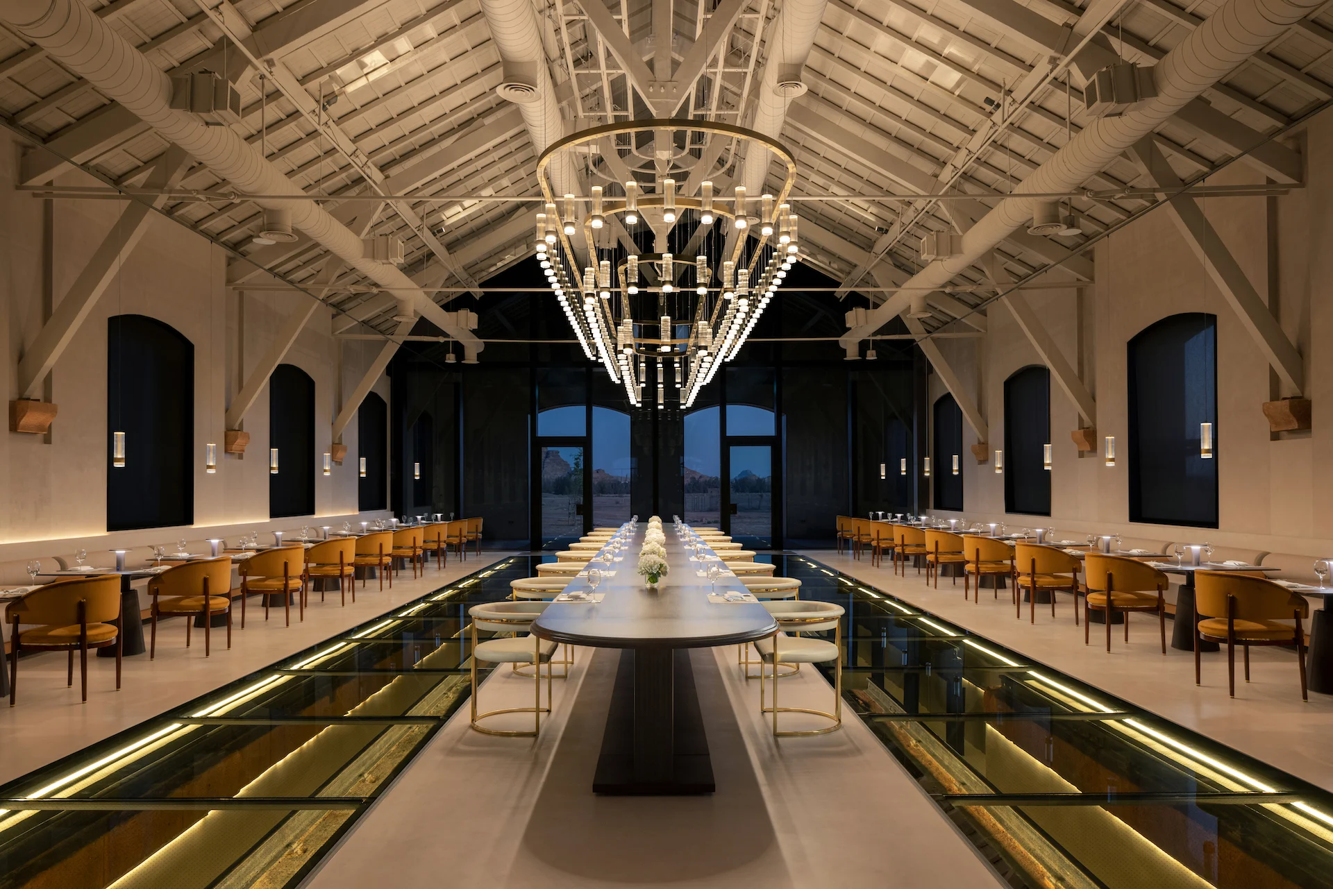 Restaurant in a restored hall: long table under a ring chandelier, glass floor panels and exposed trusses – The Chedi Hegra, AlUla, Saudi Arabia, 2024 – hotel architecture