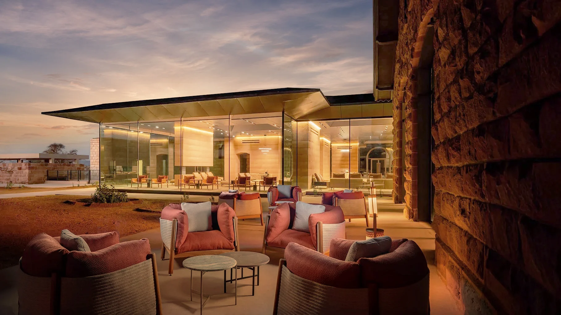 Terrace with pink armchairs at dusk in front of a glass pavilion and a rough stone wall – The Chedi Hegra, AlUla, Saudi Arabia, 2024 – hotel architecture
