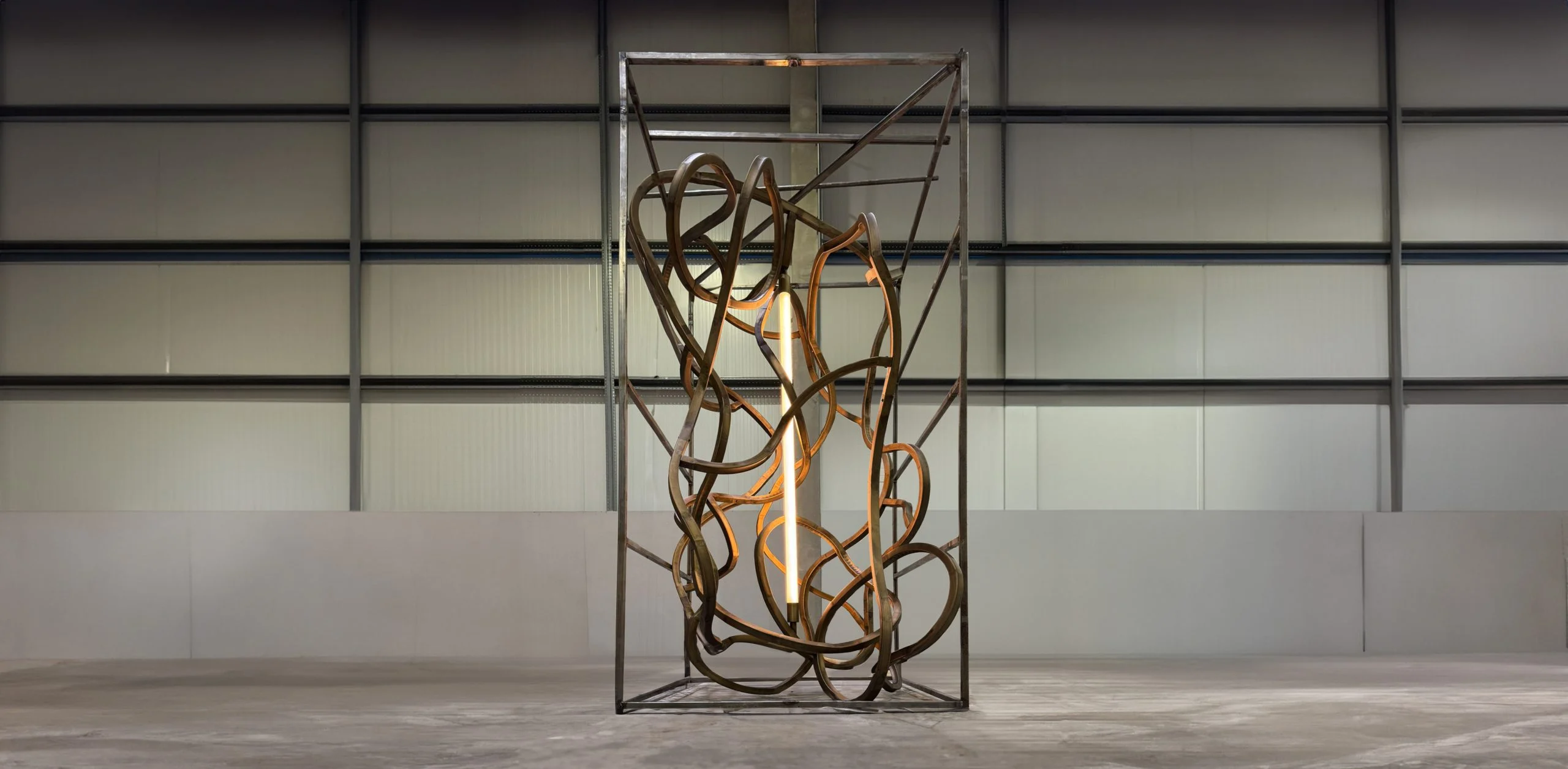 The rail chandelier upright in its tall steel frame, lit from within, in an empty hall – The Chedi Hegra Chandelier, AlUla, Saudi Arabia, 2025 – lighting design