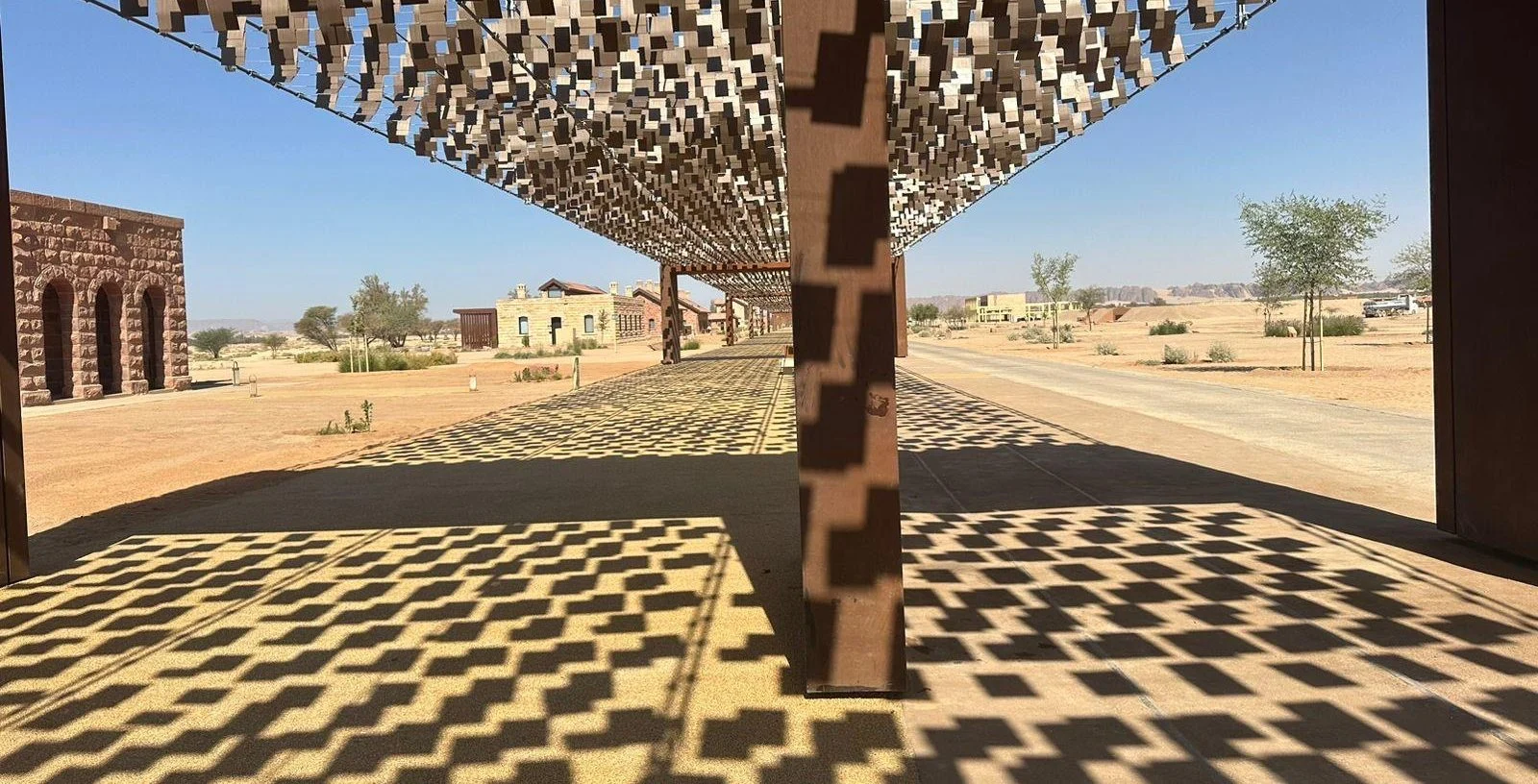 Patterned shadows cast on the ground by the kinetic canopy, with historic stone buildings and desert beyond – The Chedi Hegra Art Canopy, AlUla, Saudi Arabia, 2025 – architecture