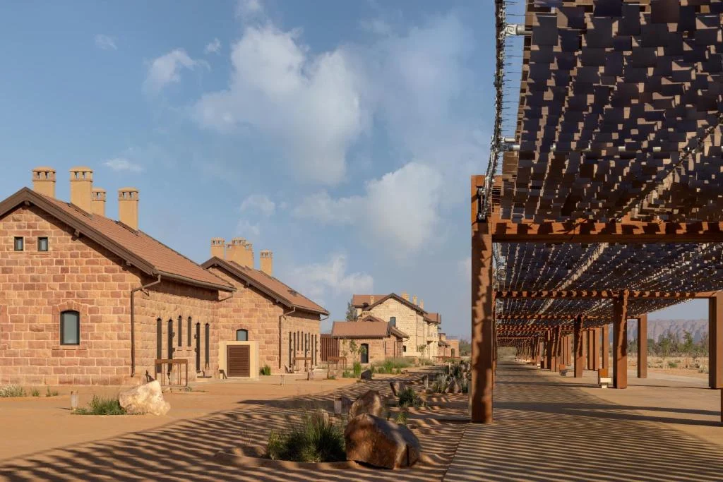 Canopy on rust-coloured posts running beside historic sandstone buildings, its shadows striping the sand – The Chedi Hegra Art Canopy, AlUla, Saudi Arabia, 2025 – architecture