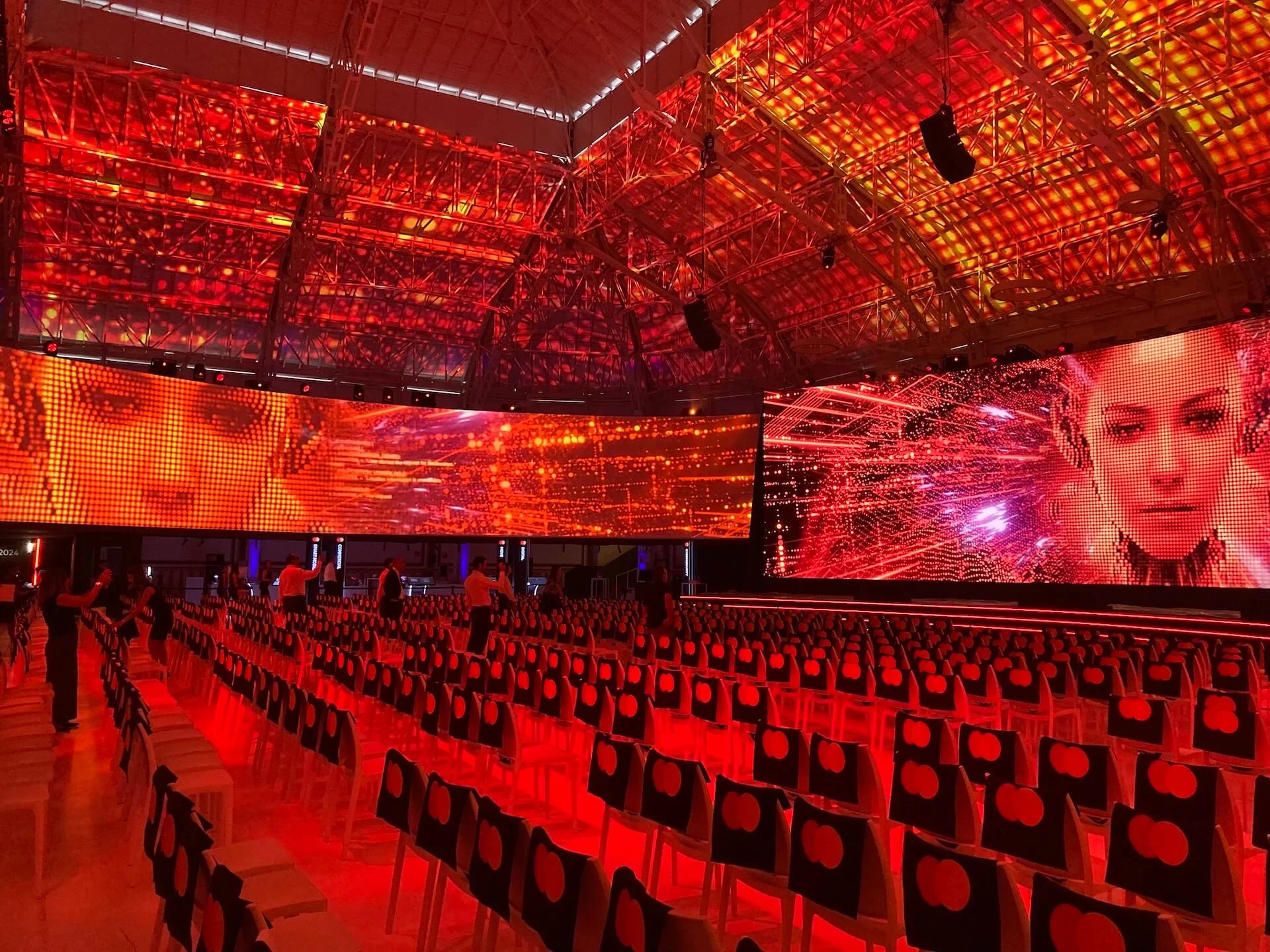 Red-lit hall with LED screens showing women's faces above rows of chairs marked with the Mastercard logo – Mastercard Innovation Forum 2024, Milan, Italy – event design