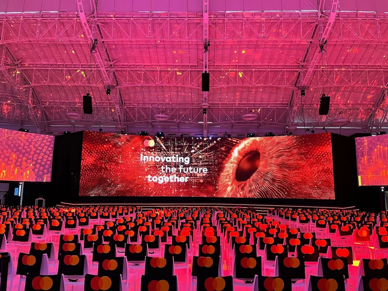 Stage screen with the words Innovating the future together over a red eye graphic, in a pink-lit hall – Mastercard Innovation Forum 2024, Milan, Italy – event design