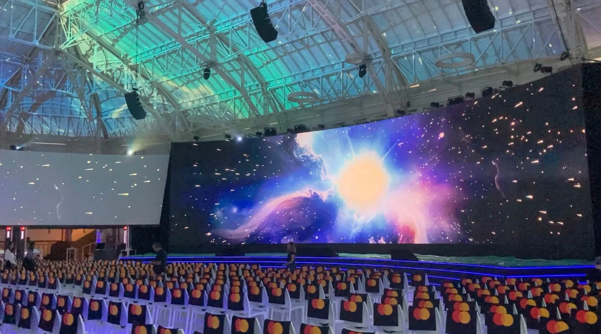Main LED screen showing a bright cosmic burst above rows of chairs, under the hall's steel roof – Mastercard Innovation Forum 2024, Milan, Italy – event design