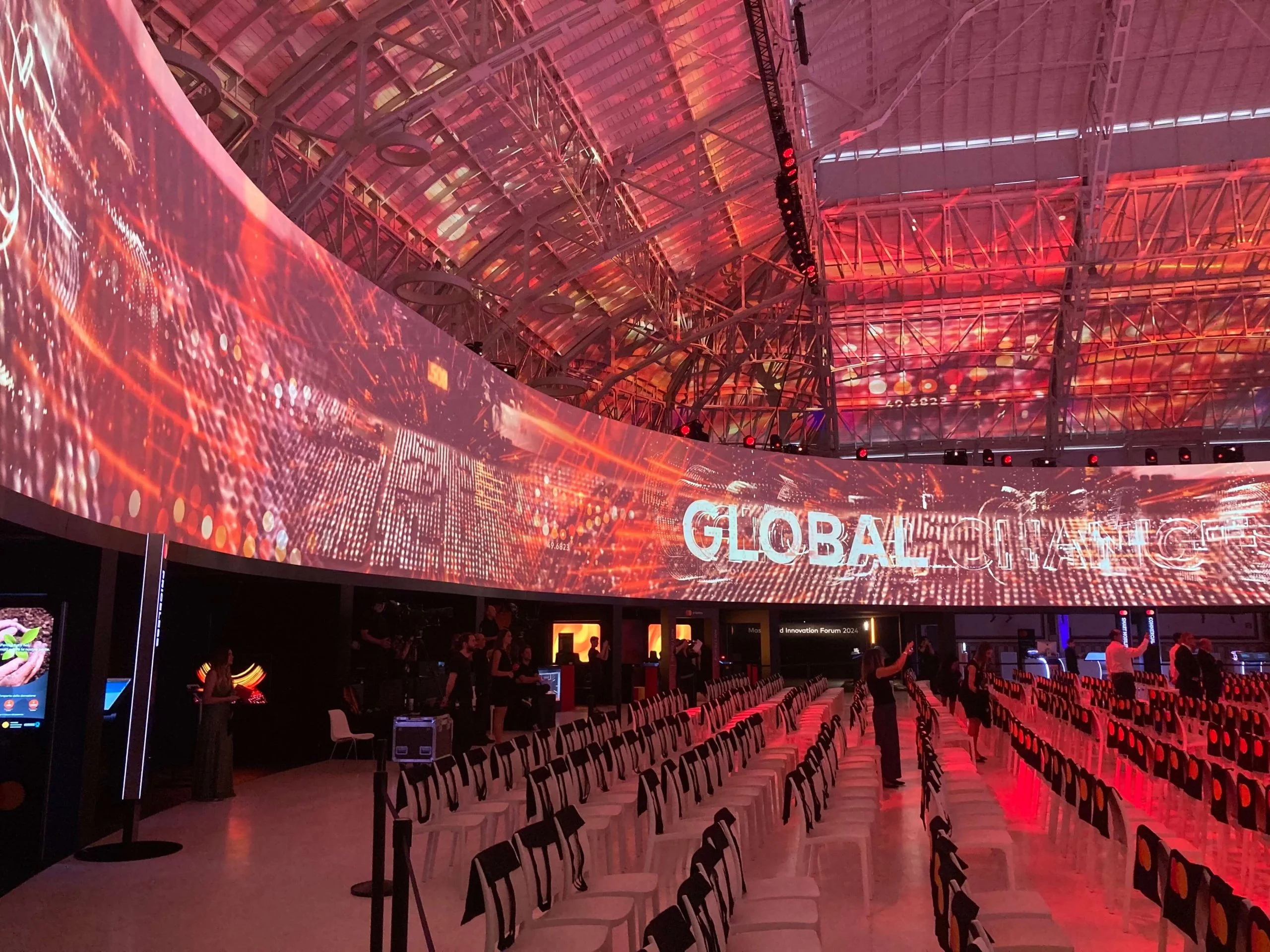 Curved LED wall wrapping the hall with red digital graphics and the word Global, chairs below – Mastercard Innovation Forum 2024, Milan, Italy – event design