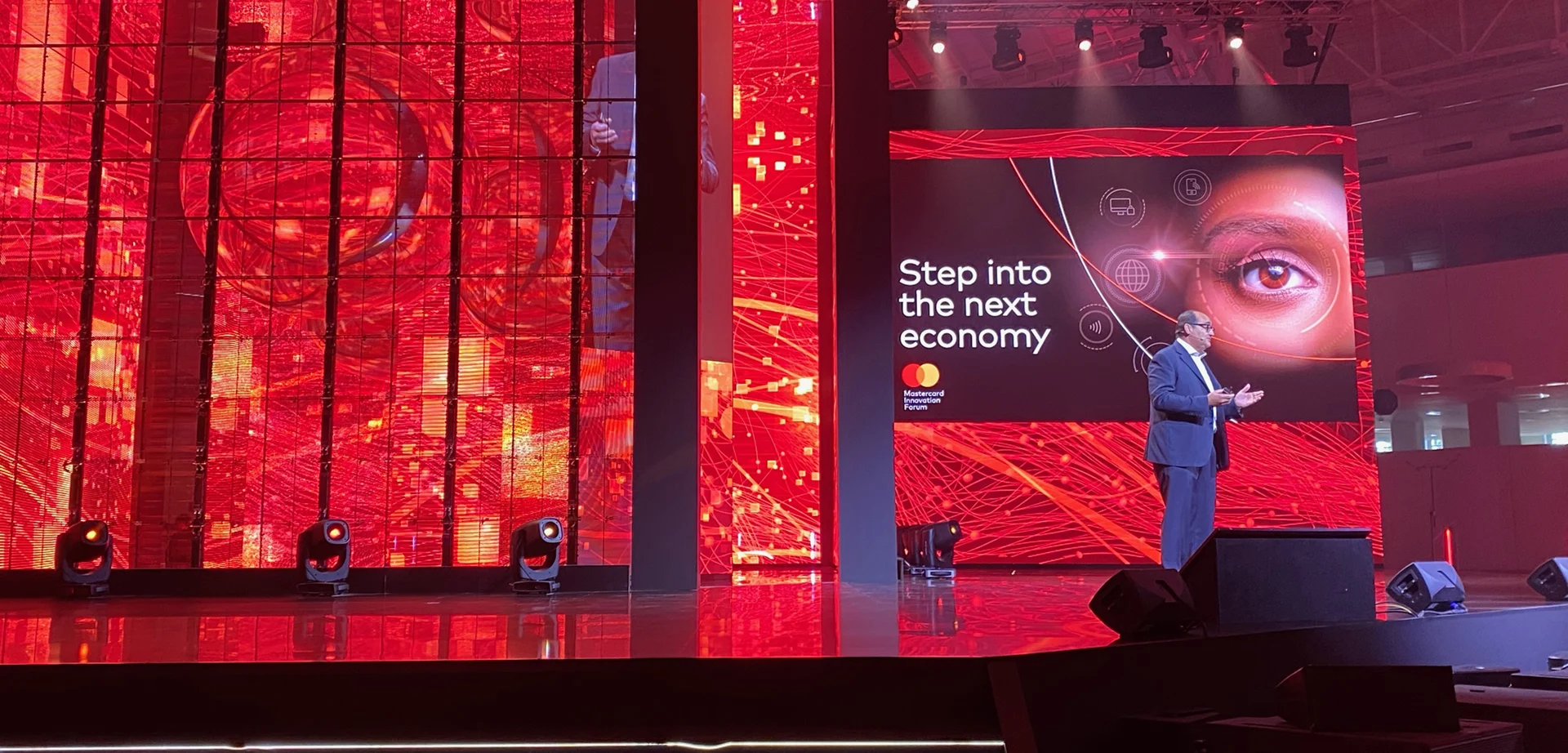 Speaker on the Mastercard Innovation Forum stage between red LED walls and a screen reading Step into the next economy – Mastercard Innovation Forum 2023, Milan, Italy – event design