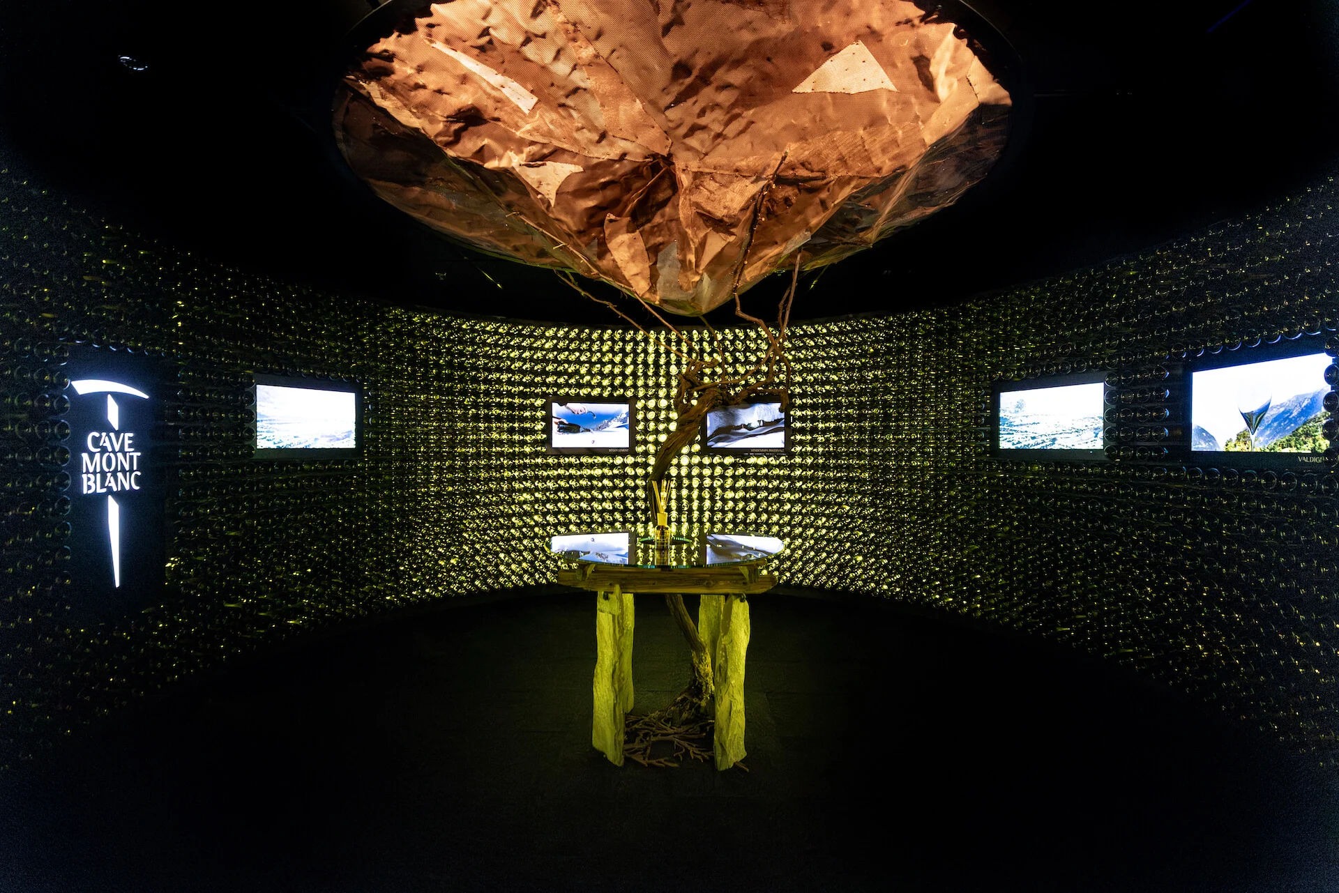 Copper-toned suspended sculpture over the vine table, with video screens set into the bottle walls – Cave Mont Blanc Winery at Skyway, Courmayeur, Italy, 2025 – interior and exhibition design
