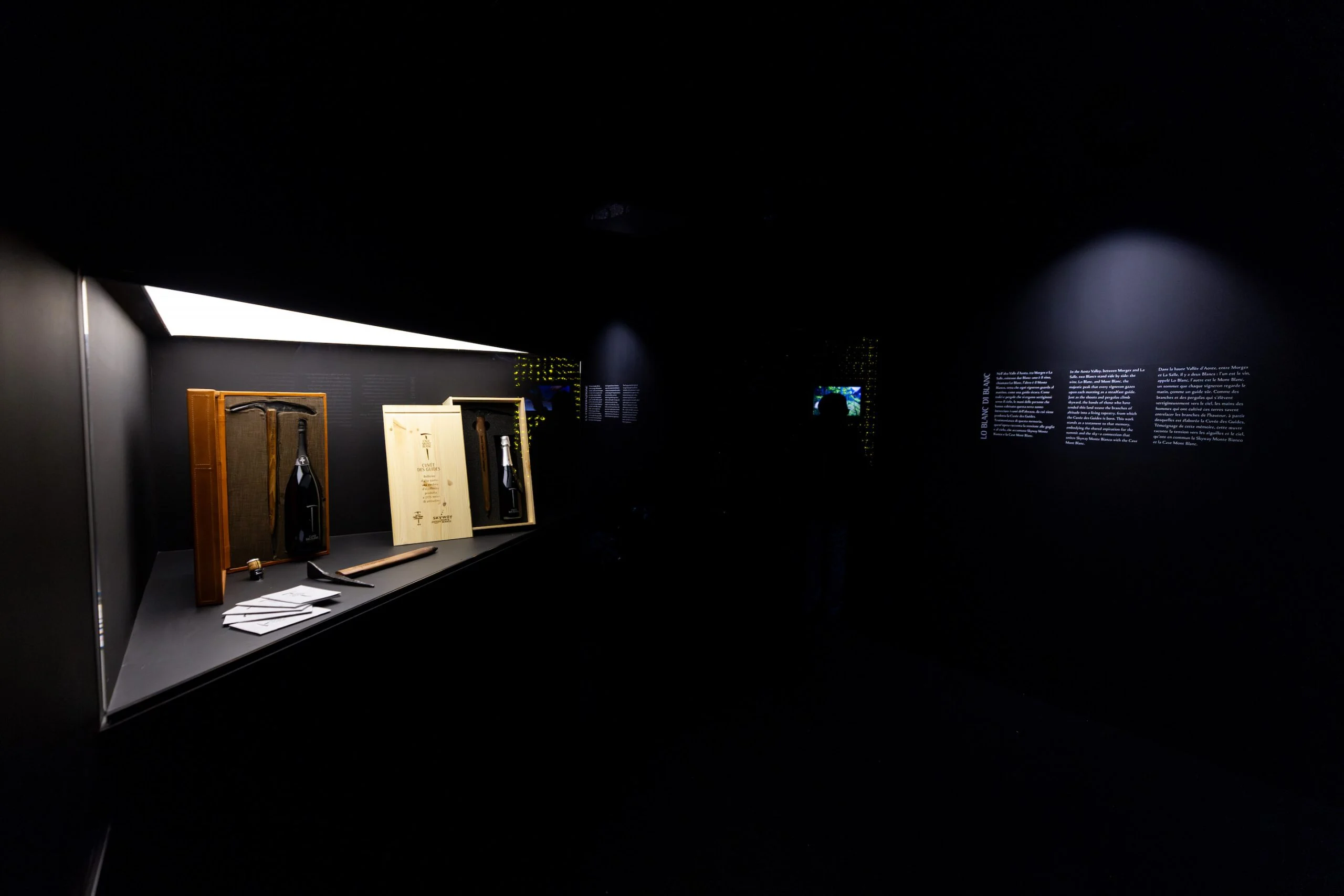 Dimly lit showcase with bottles in wooden cases and old tools, wall texts glowing in the dark room – Cave Mont Blanc Winery at Skyway, Courmayeur, Italy, 2025 – interior and exhibition design