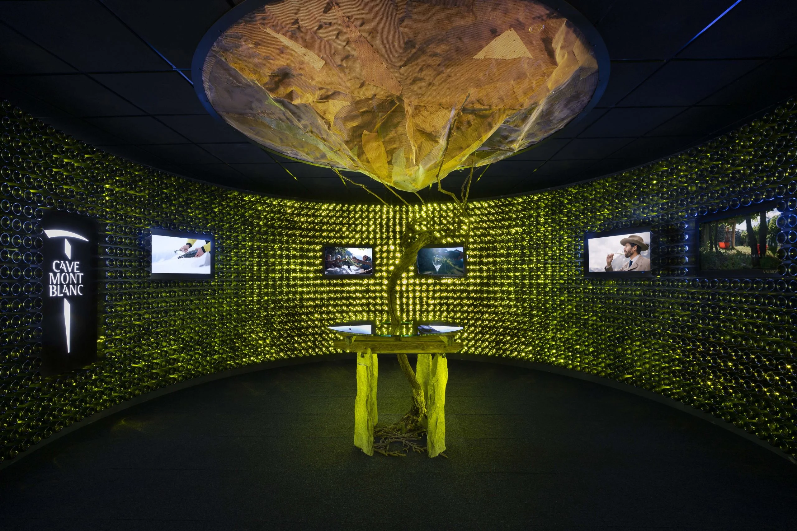 Circular room lined with backlit green bottles, a vine on a table under a suspended rock-like sculpture – Cave Mont Blanc Winery at Skyway, Courmayeur, Italy, 2025 – interior and exhibition design