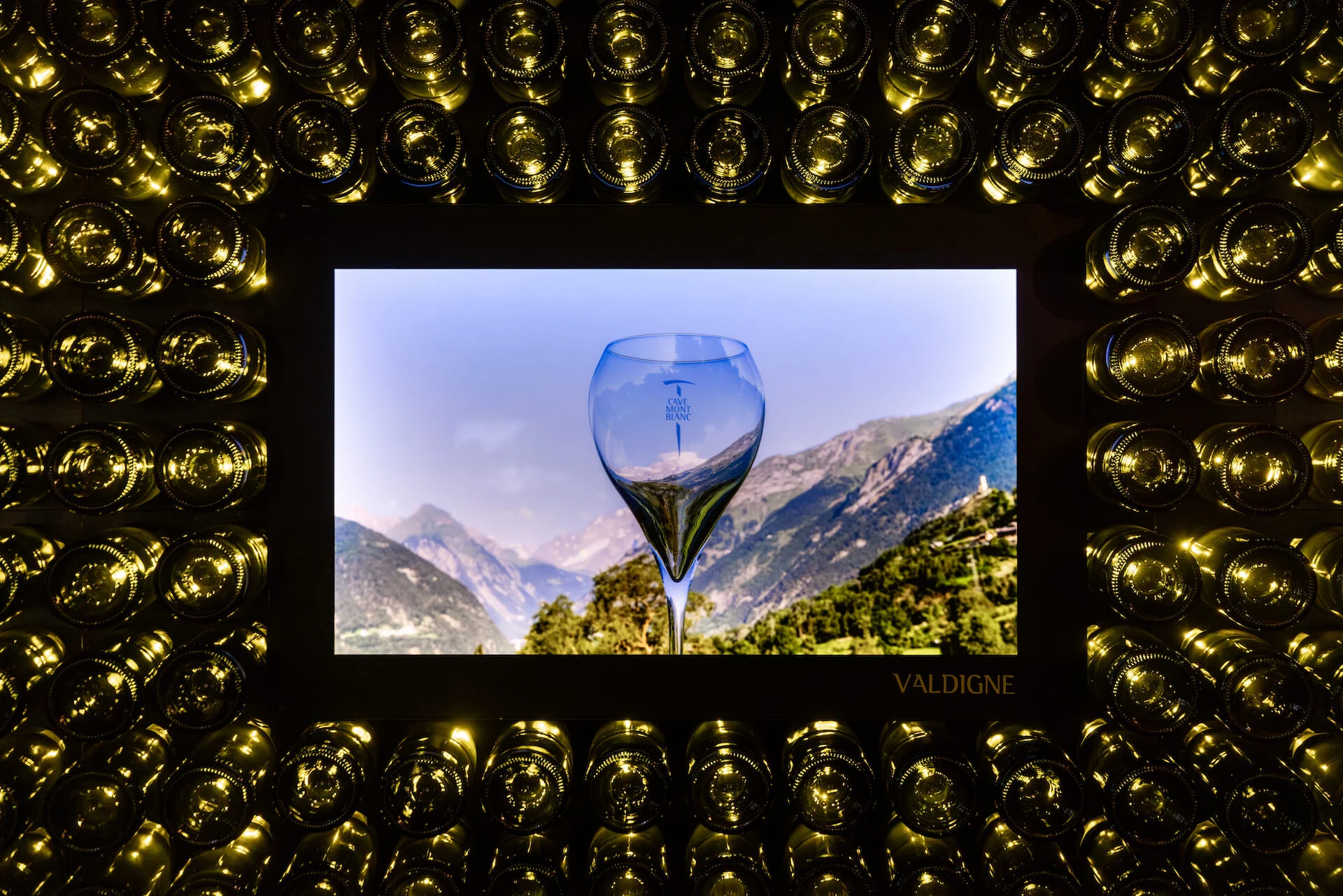 Close-up of a screen framed by bottle ends, showing a wine glass against a mountain valley – Cave Mont Blanc Winery at Skyway, Courmayeur, Italy, 2025 – interior and exhibition design