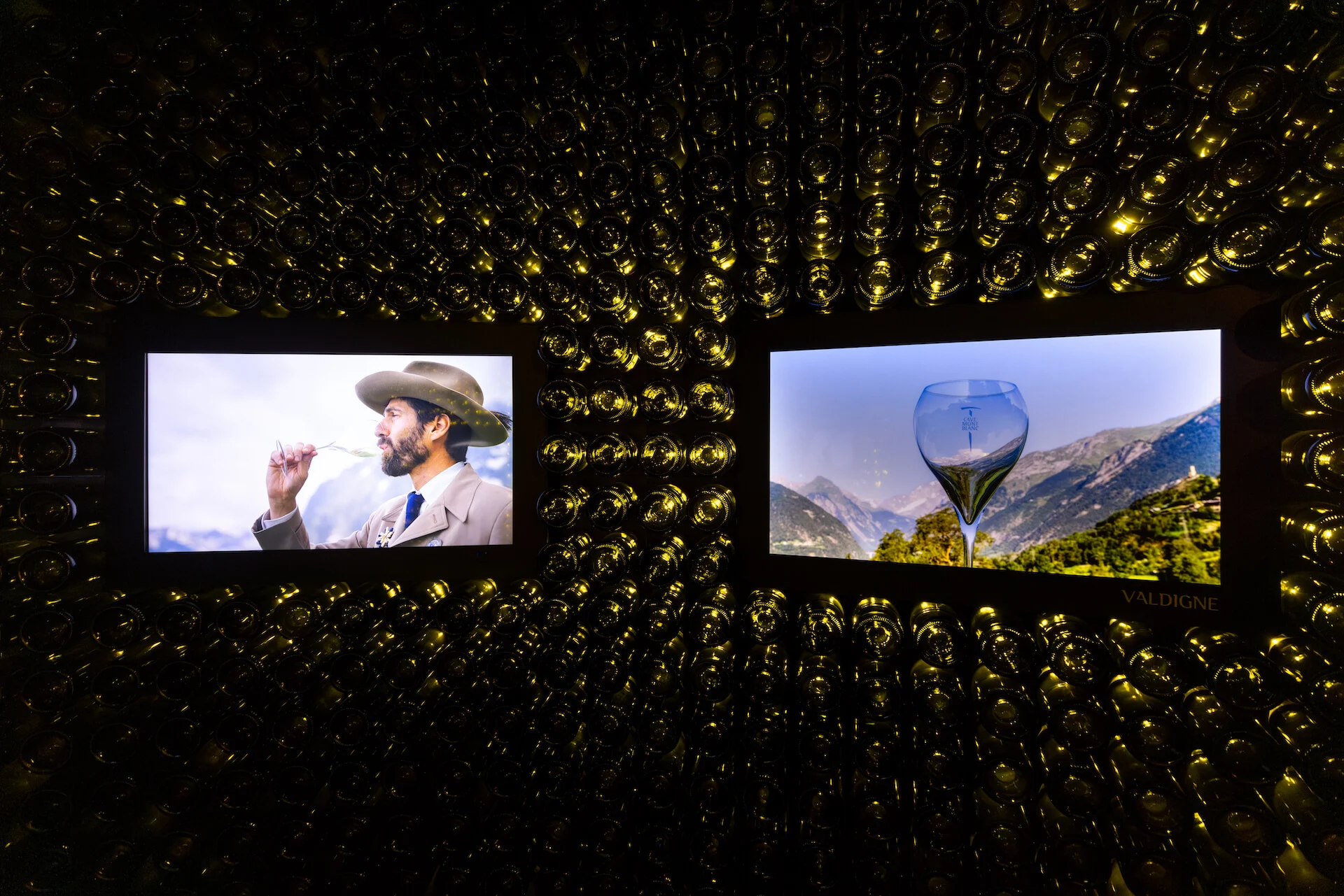 Two screens set into the bottle wall: a man tasting wine and a glass against Alpine peaks – Cave Mont Blanc Winery at Skyway, Courmayeur, Italy, 2025 – interior and exhibition design