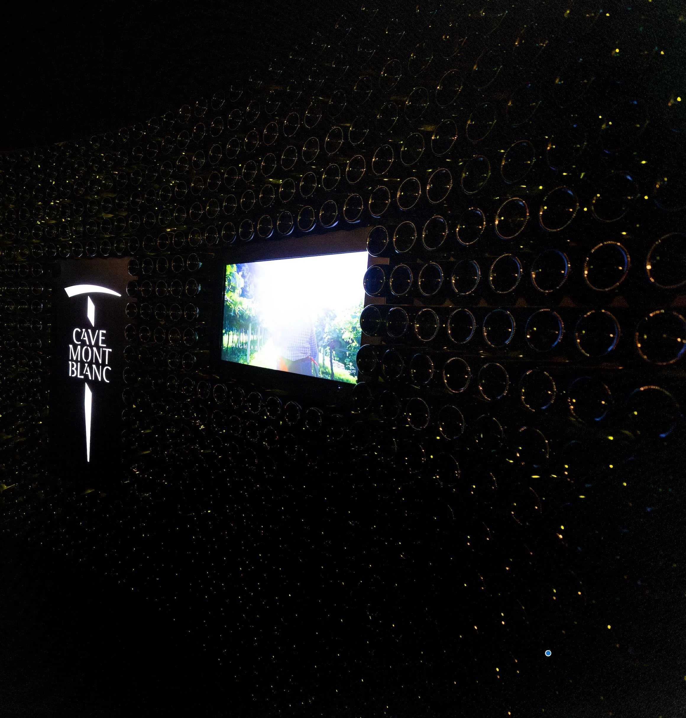 Dark wall of bottle ends with the illuminated Cave Mont Blanc logo and a screen showing vineyards – Cave Mont Blanc Winery at Skyway, Courmayeur, Italy, 2025 – interior and exhibition design