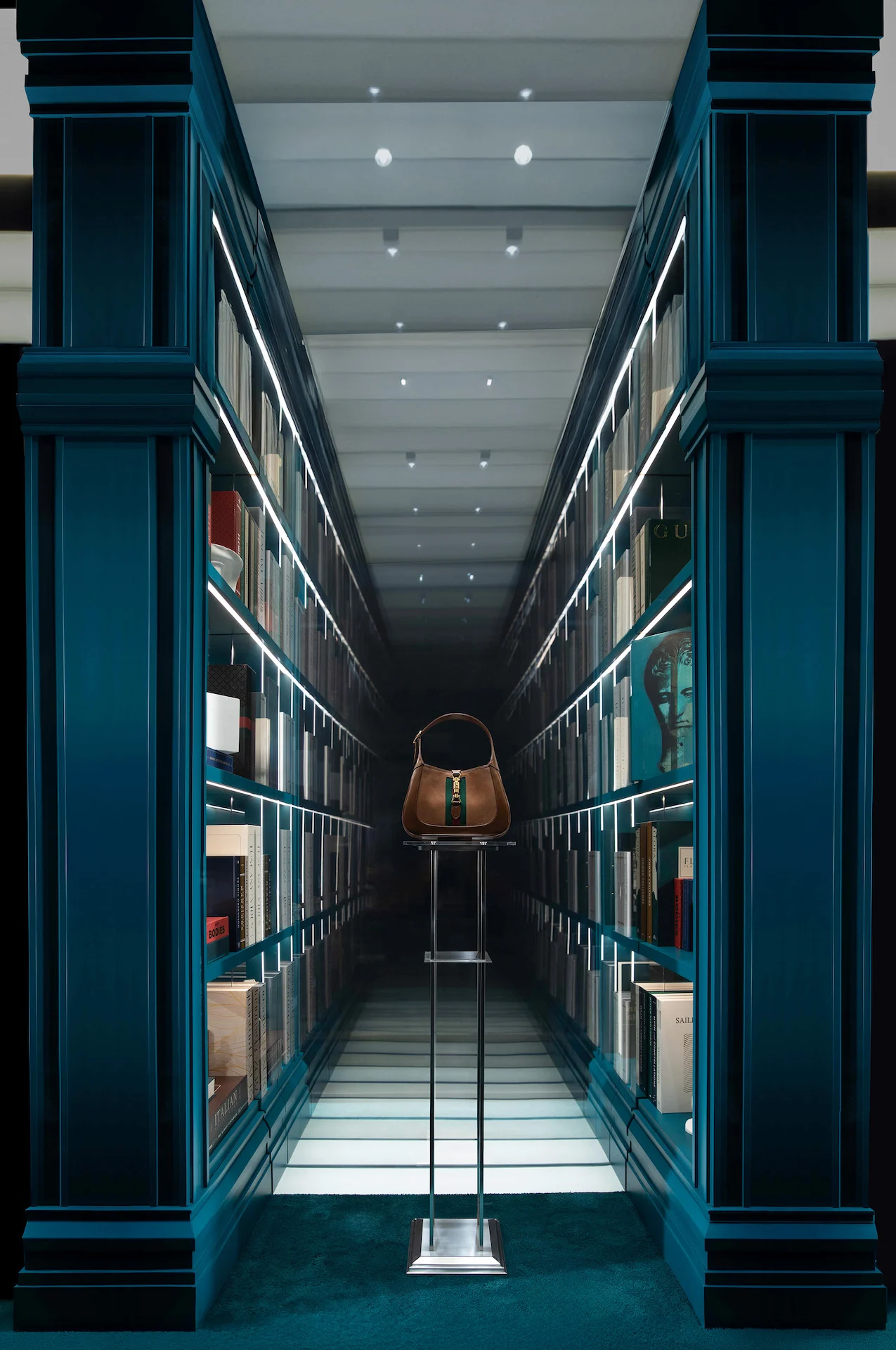 Mirrored corridor of blue-green bookcases with a Gucci handbag on a stand at its centre – Gucci Endless Narratives Window Displays, 2025 – retail design