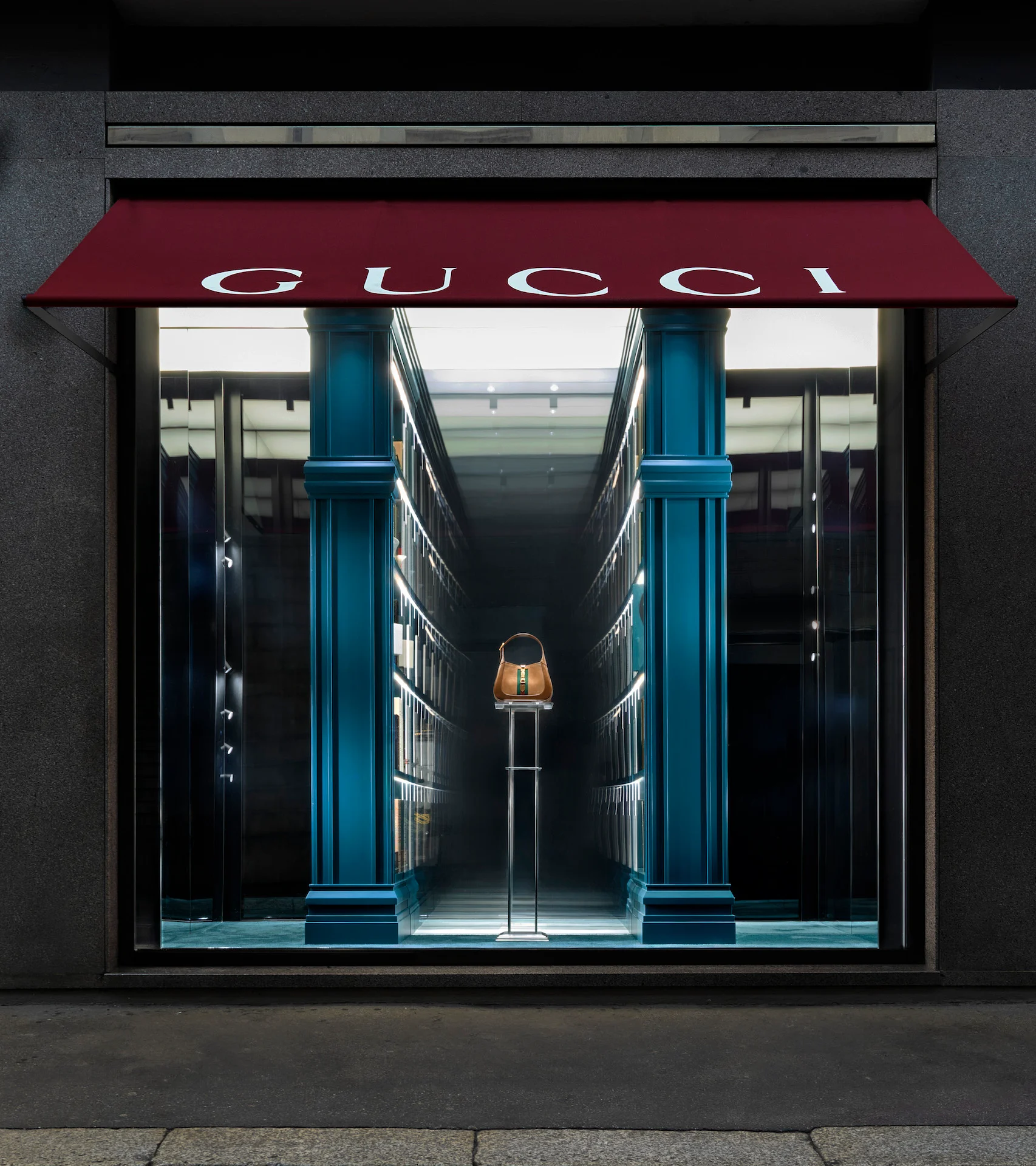Gucci window under a red awning: a single handbag at the end of an endless bookcase corridor – Gucci Endless Narratives Window Displays, 2025 – retail design