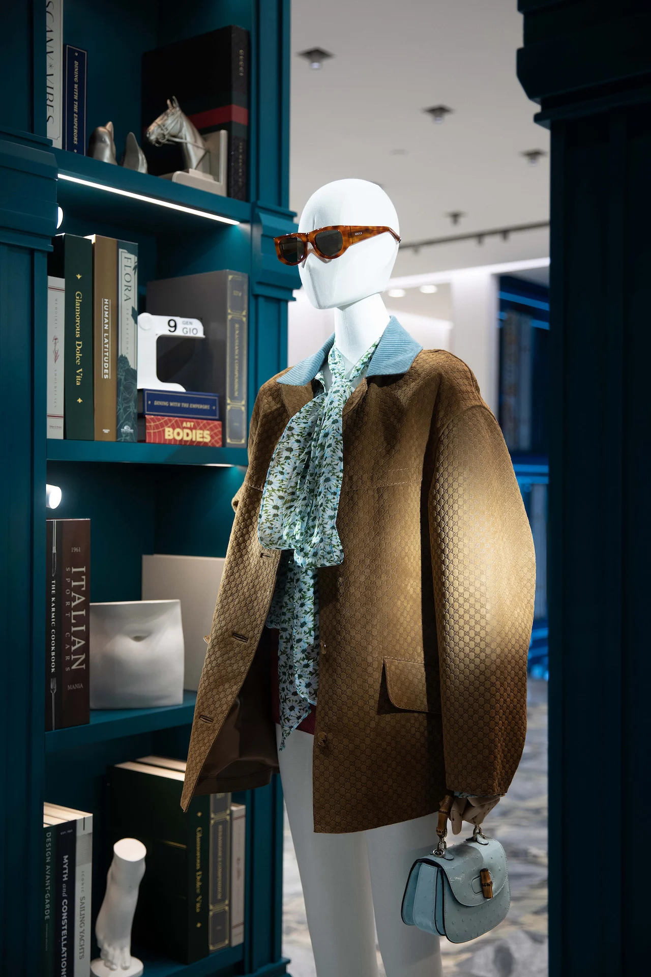 Mannequin in a suede jacket and sunglasses beside a blue-green bookcase with art books and objects – Gucci Endless Narratives Window Displays, 2025 – retail design