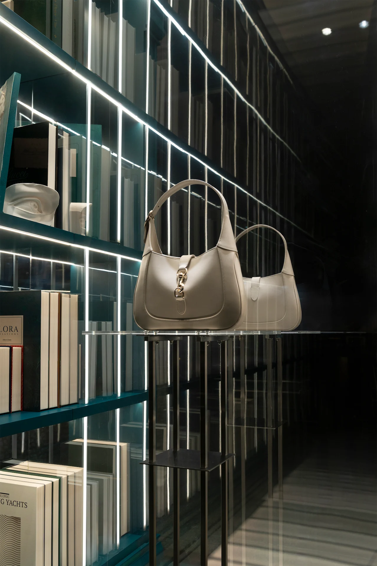 Two cream handbags on a glass shelf against mirrored bookcases lined with light strips – Gucci Endless Narratives Window Displays, 2025 – retail design