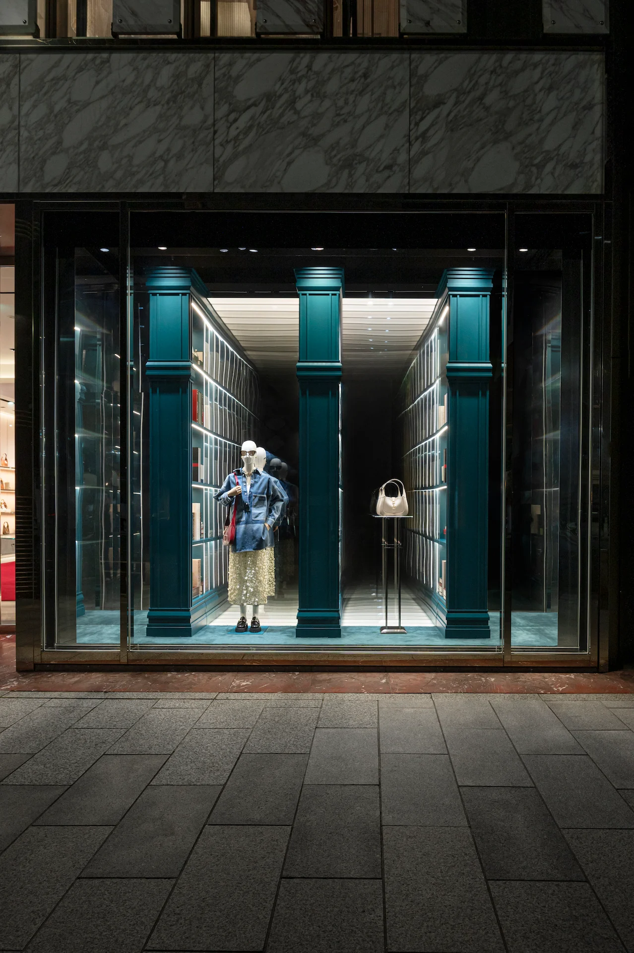 Night view of a Gucci window with a mannequin and a white bag between teal columns and bookcases – Gucci Endless Narratives Window Displays, 2025 – retail design