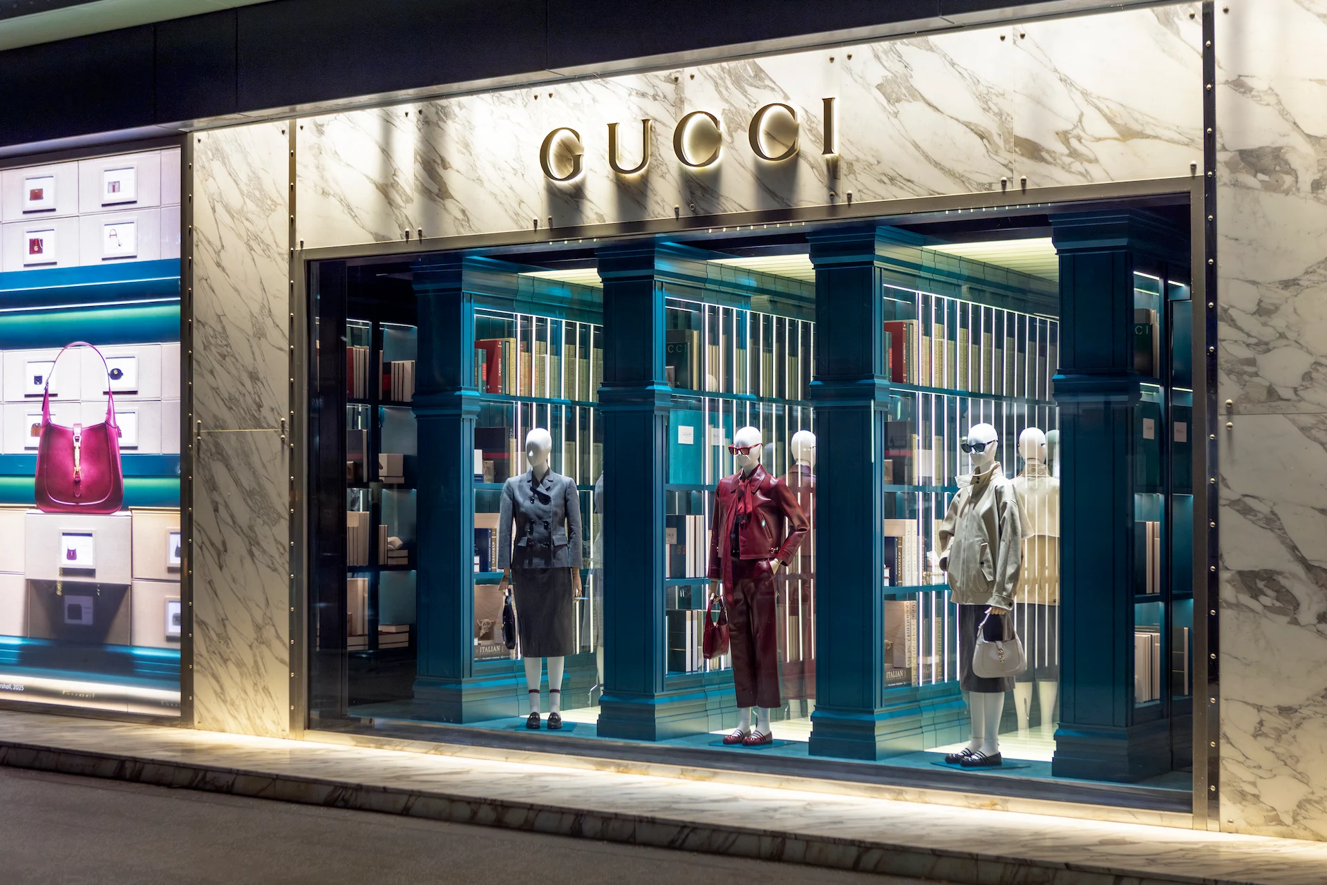 Angled view of a Gucci window with three mannequins and lit teal bookcases under a gold logo – Gucci Endless Narratives Window Displays, 2025 – retail design