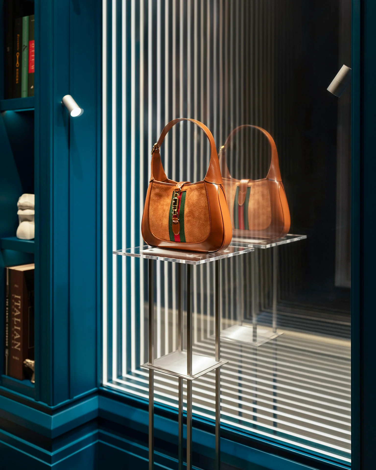Detail of a tan suede Gucci bag on an acrylic stand, reflected in a mirror lined with vertical light strips – Gucci Endless Narratives Window Displays, 2025 – retail design