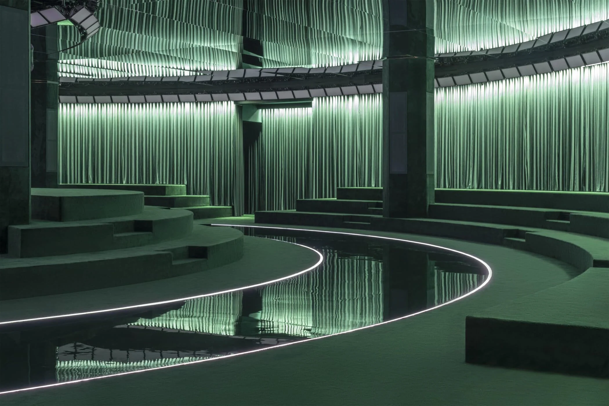 Curved runway of black mirror edged with light, winding between green velvet seating tiers – Gucci Fall/Winter 2025, Milan, Italy – fashion show set design