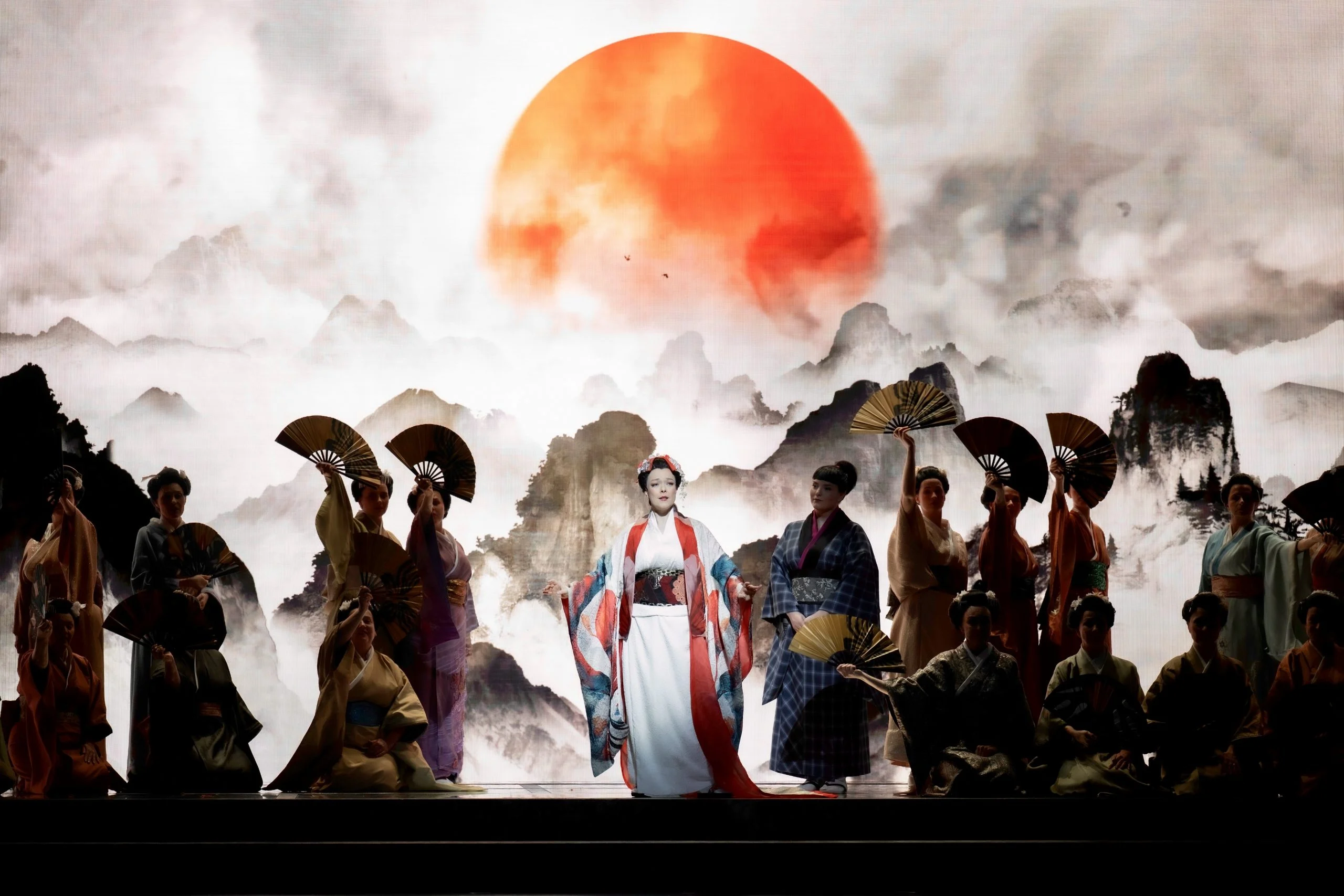 Soprano in a white and red kimono among women with fans, before a projected red sun over misty mountains – Madama Butterfly at Festspielhaus Baden-Baden, Germany, 2025 – opera set design