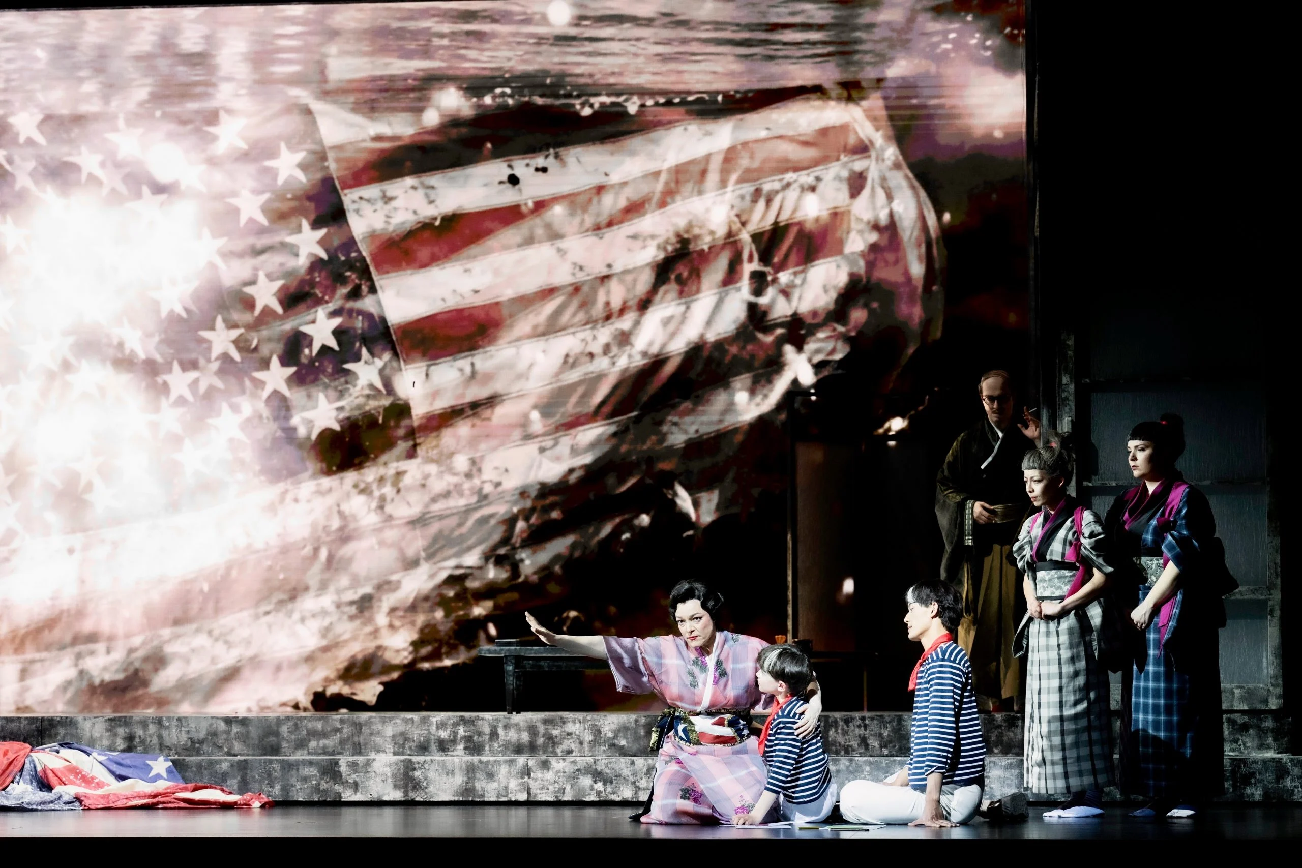 Singers in kimonos on stage before a huge projected American flag, a crumpled flag on the floor – Madama Butterfly at Festspielhaus Baden-Baden, Germany, 2025 – opera set design