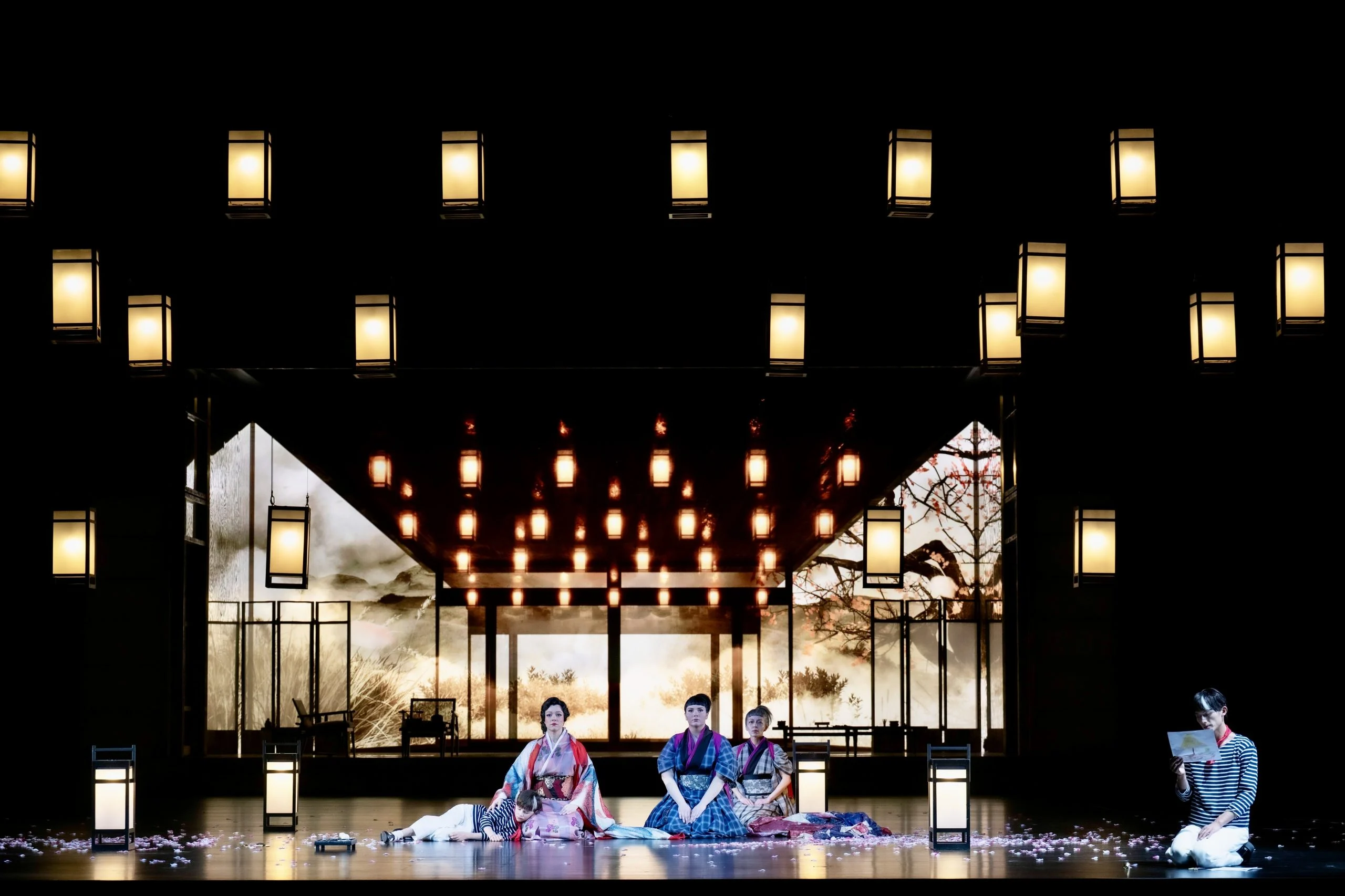 Three women kneeling on a petal-strewn stage, framed by a grid of hanging lanterns and a lit pavilion – Madama Butterfly at Festspielhaus Baden-Baden, Germany, 2025 – opera set design