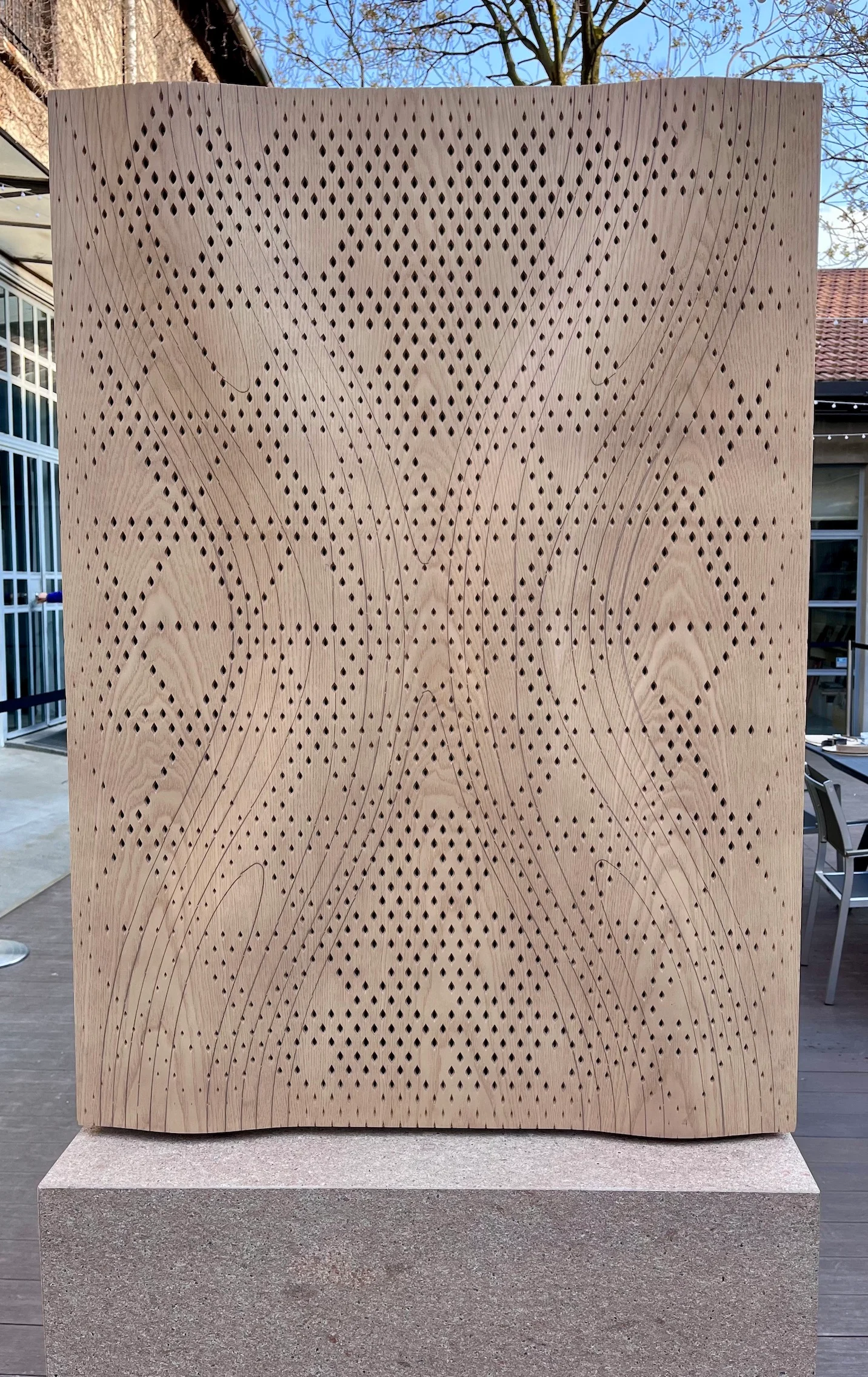 Front view of a wavy perforated wooden acoustic panel on a stone plinth in a courtyard – Strong by Form Woodflow-Skin Panels, Milan, Italy, 2025 – product design