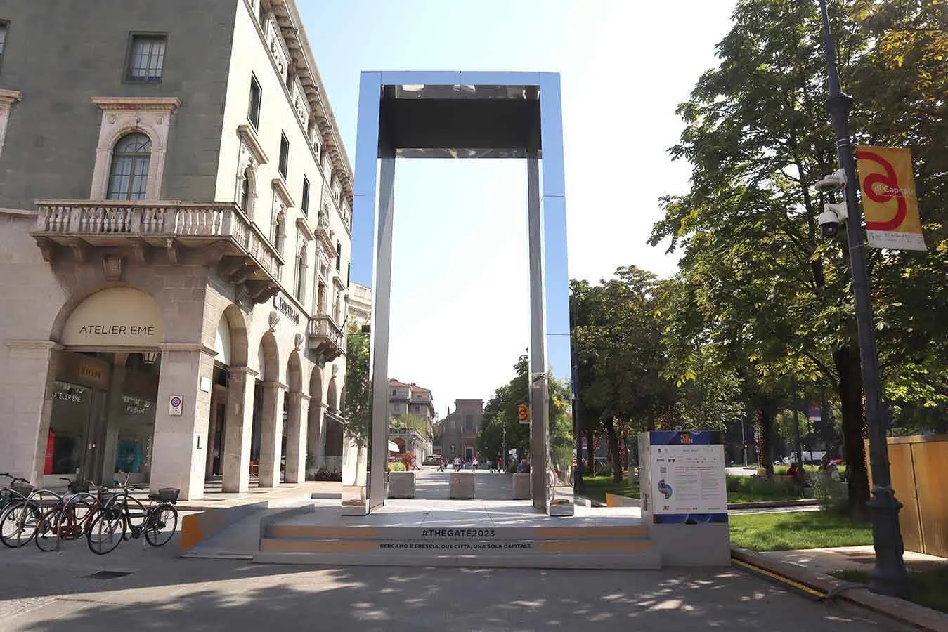 Mirror-clad THE GATE portal on a raised platform in a tree-lined city square on a sunny day – THE GATE 2023, Bergamo and Brescia, Italy – installation design