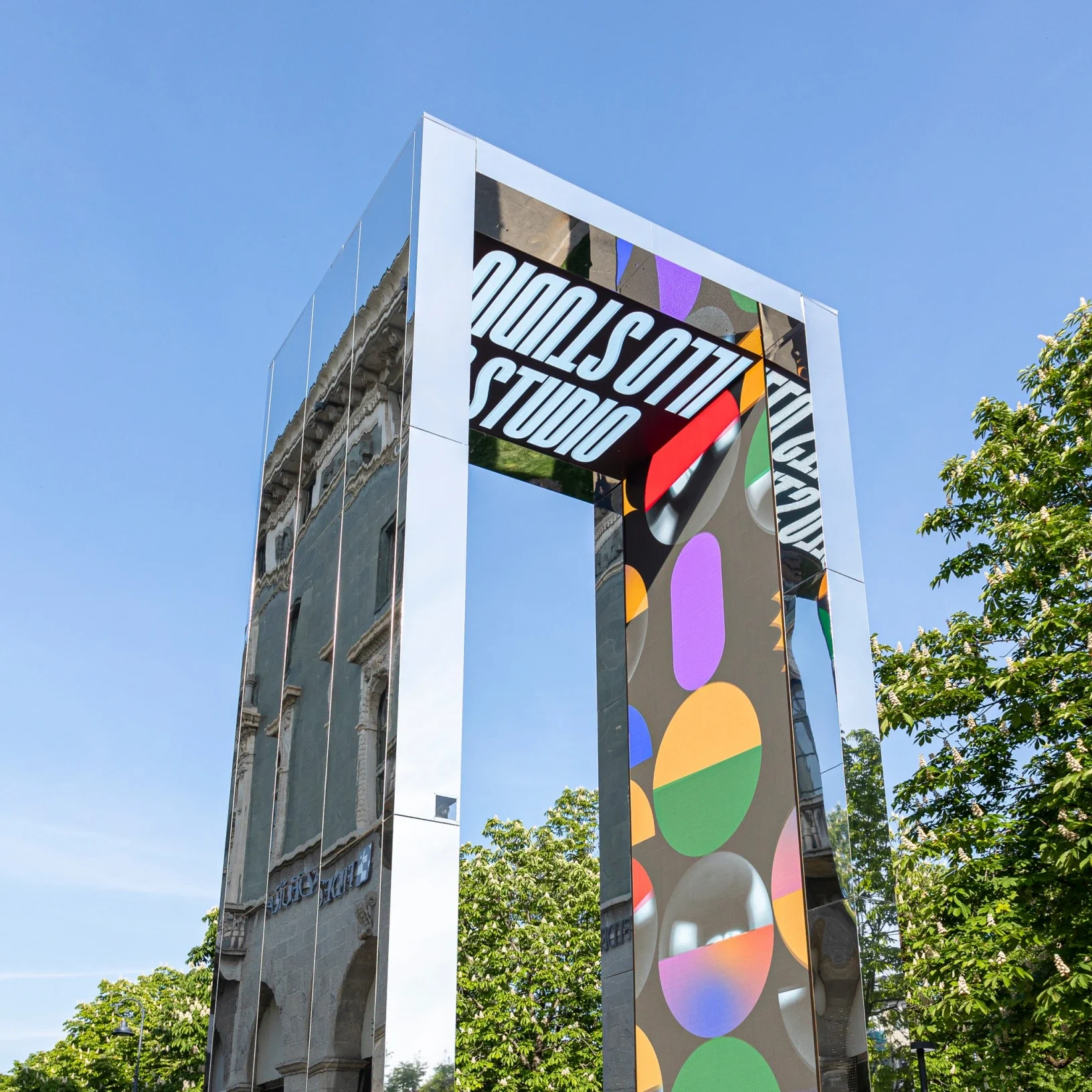 Low-angle view of the mirrored portal with colourful LED graphics and Studio lettering against a blue sky – THE GATE 2023, Bergamo and Brescia, Italy – installation design