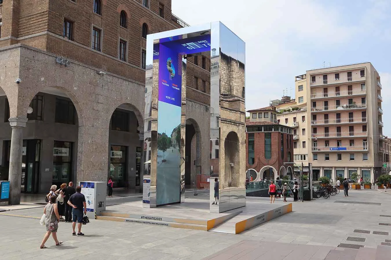 Mirror-clad portal reflecting arcaded buildings, its inner LED screen showing video as people walk past – THE GATE 2023, Bergamo and Brescia, Italy – installation design