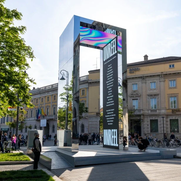 Mirrored portal with an iridescent LED lintel and a tall UNREEL screen, people gathered around it – THE GATE 2023, Bergamo and Brescia, Italy – installation design