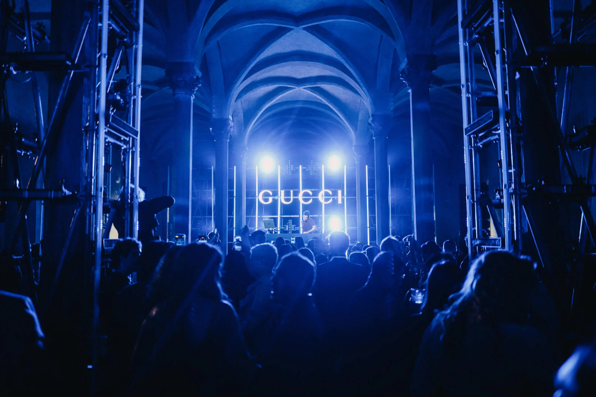 Crowd in blue light before a lit GUCCI sign and bright spotlights, scaffolding at the sides – Gucci Cruise 2026 Afterparty, Florence, Italy, 2025 – event design