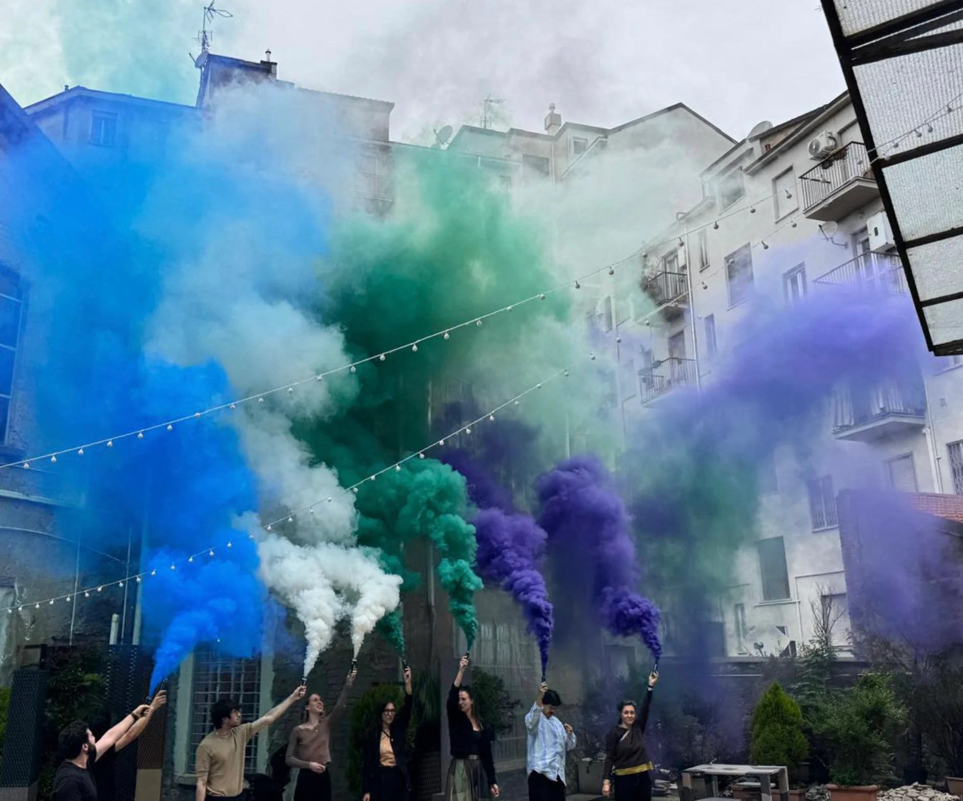 People holding blue, white, green and purple smoke flares in a courtyard between city buildings – Cesare Cremonini Live25, Italy, 2025 – concert stage design