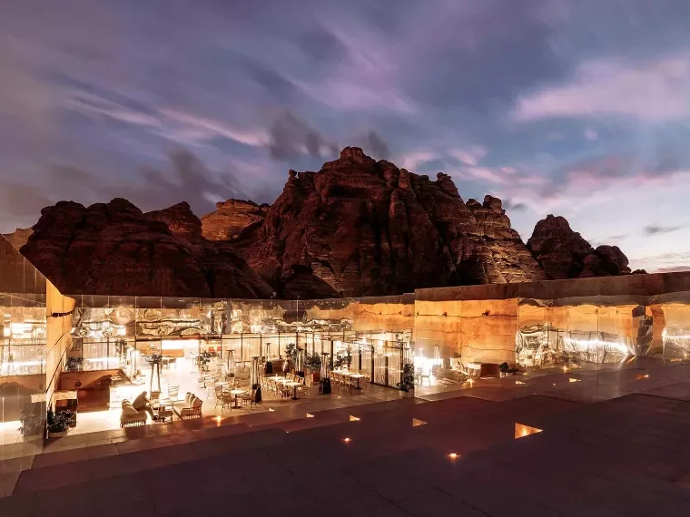 Terrace with rattan chairs and marble tables with lamps, glass wall and reflected cliffs behind – Maraya Concert Hall, AlUla, Saudi Arabia, 2019 – architecture