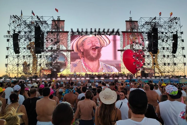 Beach crowd at sunset facing the stage, the screen showing the singer in a straw hat, flanked by a red heart and mirror ball – Jova Beach Party 2022, Italy – concert stage design