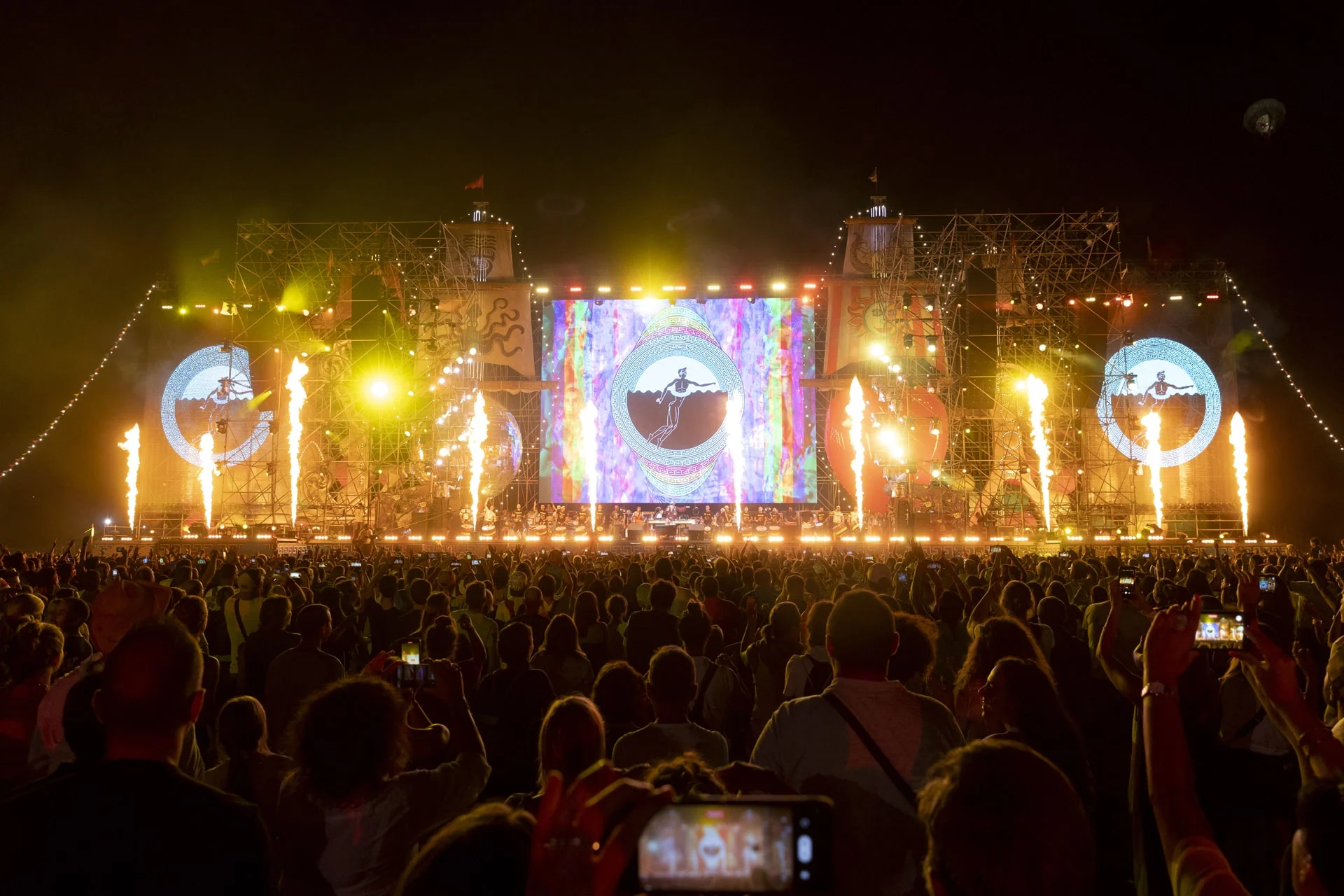 Flame jets shooting up along the stage at night, psychedelic visuals on the central and circular side screens – Jova Beach Party 2022, Italy – concert stage design