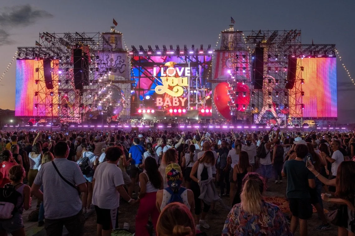 Crowd at dusk facing the illuminated stage, with "I Love You Baby" on the central screen and colourful side screens – Jova Beach Party 2022, Italy – concert stage design